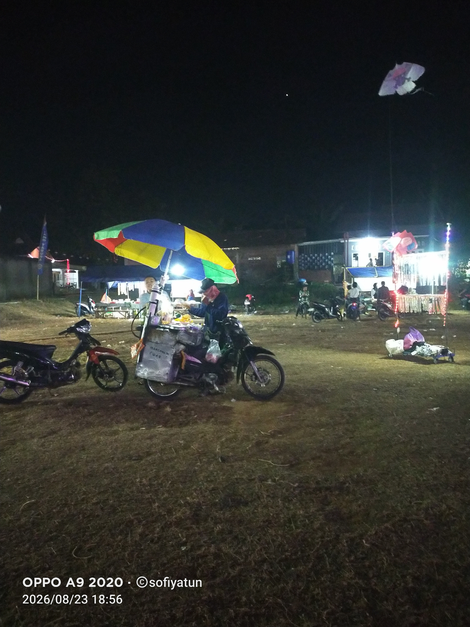 The image depicts a nighttime scene in what appears to be an outdoor market or gathering area in Cluwak, Indonesia. In the foreground, a motorcycle is parked. To its right, a food cart with a colorful umbrella is set up, attended by a person wearing a blue jacket and a dark cap. The cart displays some items, and there are plastic bags on the back of the motorcycle.
In the middle ground, several other motorcycles are parked, and a few people are visible near a building with lights on. A tall, thin pole with colorful lights extends upwards on the right side of the image. In the background, there are buildings and trees, and the sky is dark. A large, decorative kite, possibly shaped like a butterfly, is flying high in the sky on the right side of the image. The ground is covered in dry grass. The timestamp "2026/08/23 18:56" and a copyright notice are visible at the bottom left of the image.