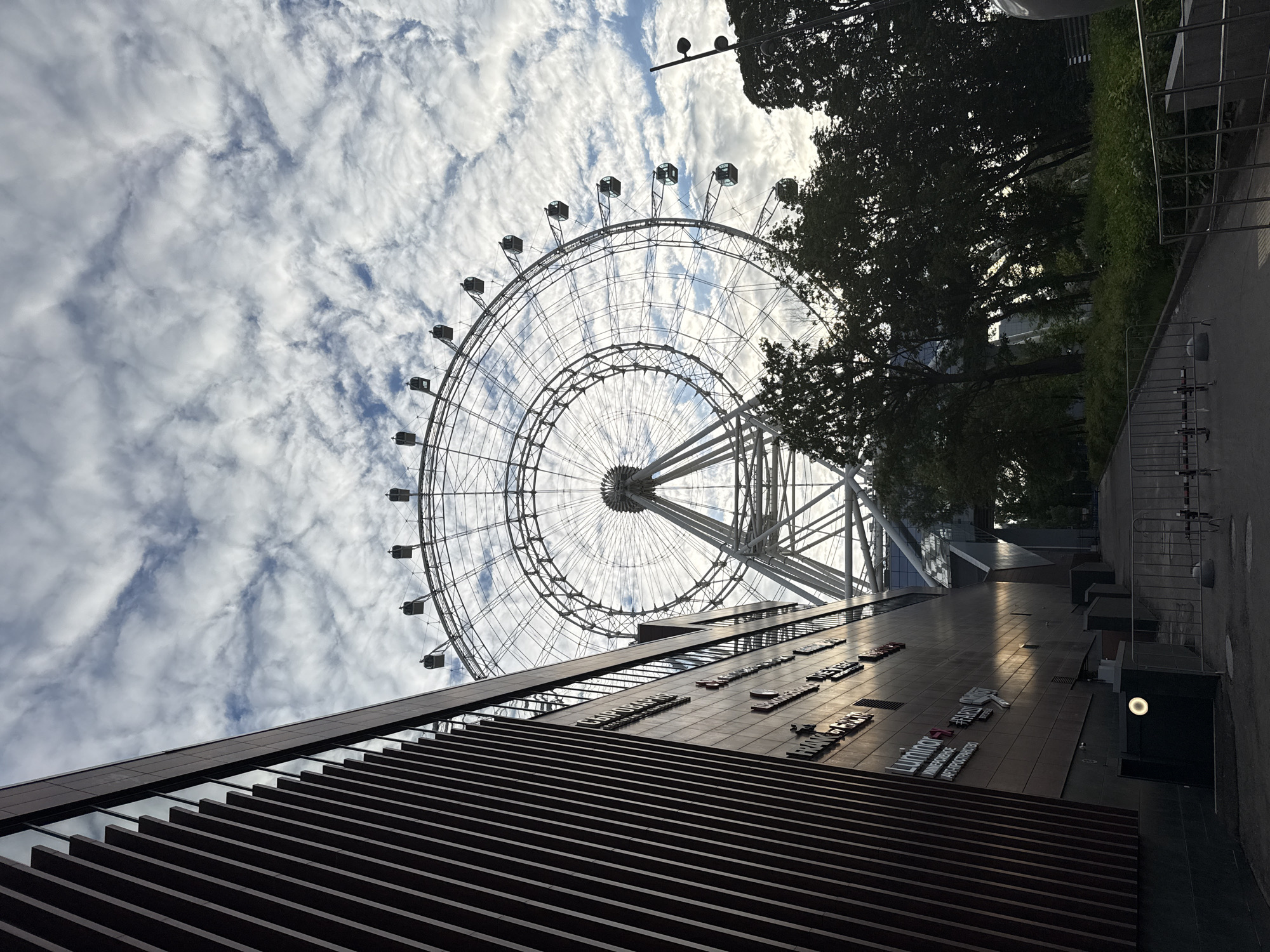 An architectural and landscape photograph taken from a low-angle perspective, looking upwards at a Ferris wheel and a modern building against a cloudy sky. The scene is set in an urban environment, identified as Moscow, Russia.
The primary subject is a large Ferris wheel, its intricate metal framework and numerous passenger gondolas dominating the upper portion of the frame. It is silhouetted against a bright, partly cloudy sky filled with textured white and grey clouds. To the right of the Ferris wheel, the dark, dense foliage of trees is visible, also silhouetted against the bright sky.
In the lower part of the image, a contemporary building facade extends from the bottom, receding towards the middle distance. The lowest section features dark, horizontal slatted panels. Above this, a tiled section of the building displays several pieces of text, including "LUMINOR," "FRANK & MARIE," "PARFUM," and "КОММЕРЧЕСКИЕ ПАРТНЁРЫ ОРГАНАЙЗЕРА" (Commercial Partners of the Organizer). The building appears to have modern design elements. Further to the right, a paved area with metal barriers or fences, along with some street lighting and infrastructure, is partially visible.
The time of day appears to be daylight, possibly early morning or late afternoon, given the bright sky and the way elements like the Ferris wheel and trees are backlit, creating strong contrasts and shadows. No people are visible, and there is no specific activity depicted beyond the static structures of the amusement ride and building. The weather is partly cloudy. The overall impression is that of a modern leisure or commercial area.