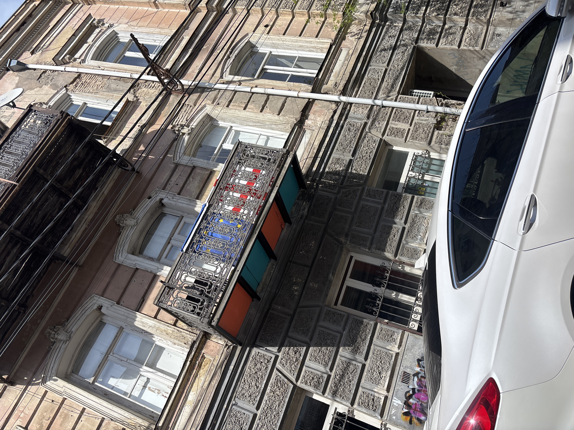 Exterior shot of buildings and a car in Tbilisi, Georgia. Multi-story buildings with varying architectural styles, window designs, and balconies. One balcony is colorfully decorated with red, white, blue, and orange panels. The buildings are a mix of pink and gray. Electrical wires run along the facade. A white car is parked partially obscuring one building's facade. Graffiti is visible near the car. The scene appears to be sunlit.