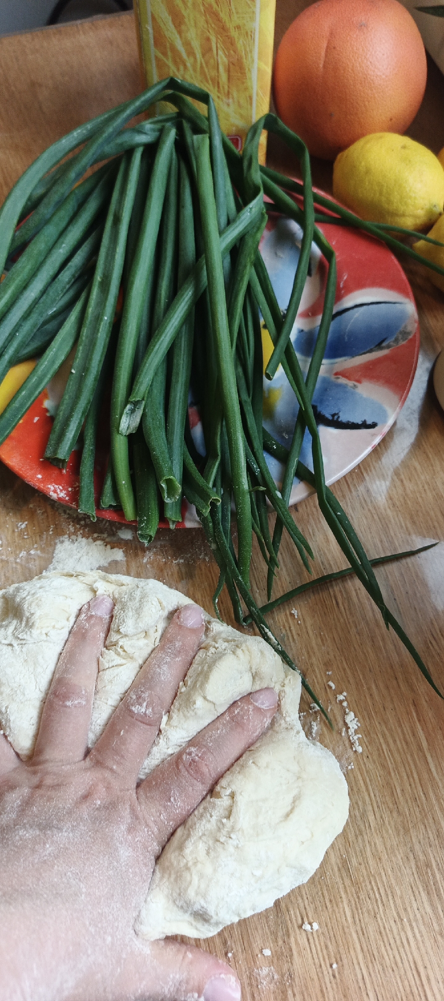 A human hand with manicured fingernails is pressed into a mound of light-colored dough on a wooden surface. The dough is dusted with white powder, likely flour. To the upper right of the dough, a pile of long, green stalks of green onions rests on a colorful, circular plate with a floral design. In the background, to the left of the green onions, is a portion of a yellow and white striped box. Further to the right, a large grapefruit and a lemon are visible. The lighting is bright, casting some shadows on the wooden surface. The scene suggests food preparation.