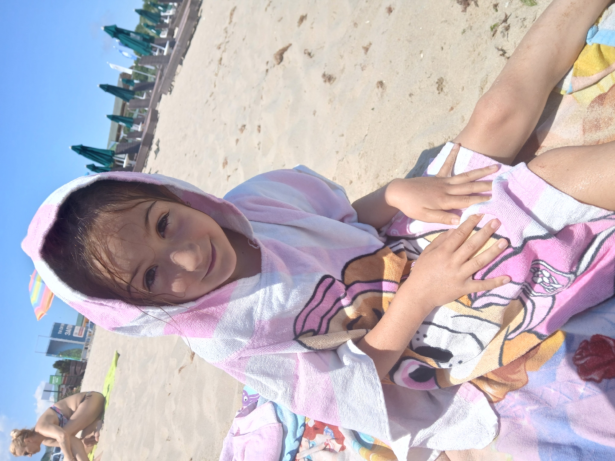 A young girl is lying on a sandy beach, wrapped in a hooded towel with pink and white stripes and a Paw Patrol character print. She is smiling at the camera, her face close up. Her hands are resting on the towel. In the background, there are beach umbrellas and loungers, suggesting a developed beach area. Further back, there are some signs, one of which appears to have Cyrillic text. The sky is blue and clear, indicating a sunny day, likely in the afternoon given the brightness. Another person, an adult woman in a bikini, is visible in the distance, crouching on the sand. The overall scene conveys a relaxed and cheerful atmosphere of a day at the beach.