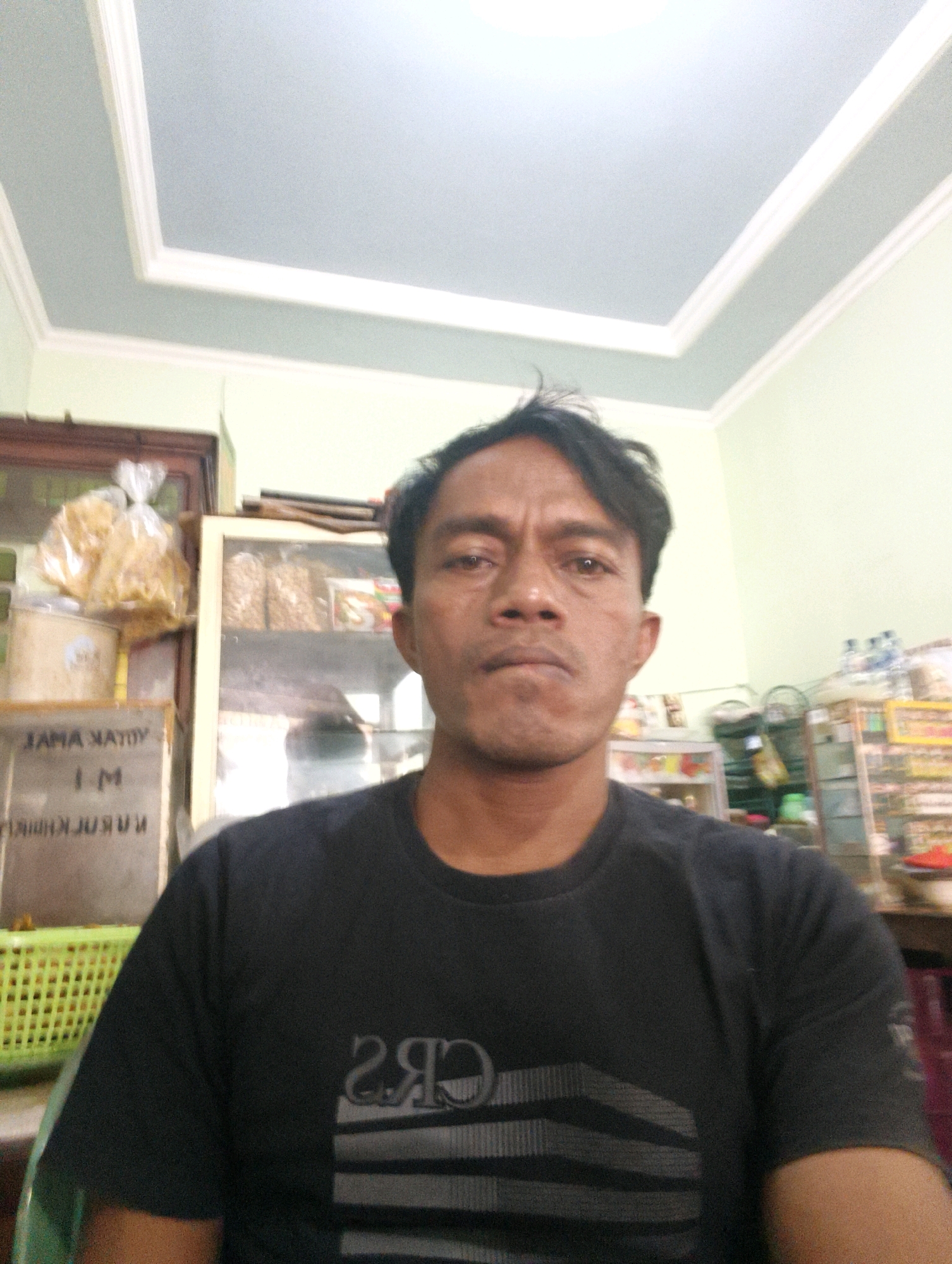 A man in a black t-shirt is posing for a selfie in front of a display shelf. He is looking directly at the camera with a neutral expression. The background shows a room with shelves stocked with various items, including snacks and bottles. There is a green basket to the left of the man. The ceiling has a decorative molding. The man's t-shirt has a graphic design on it. The overall environment appears to be a small shop or convenience store.