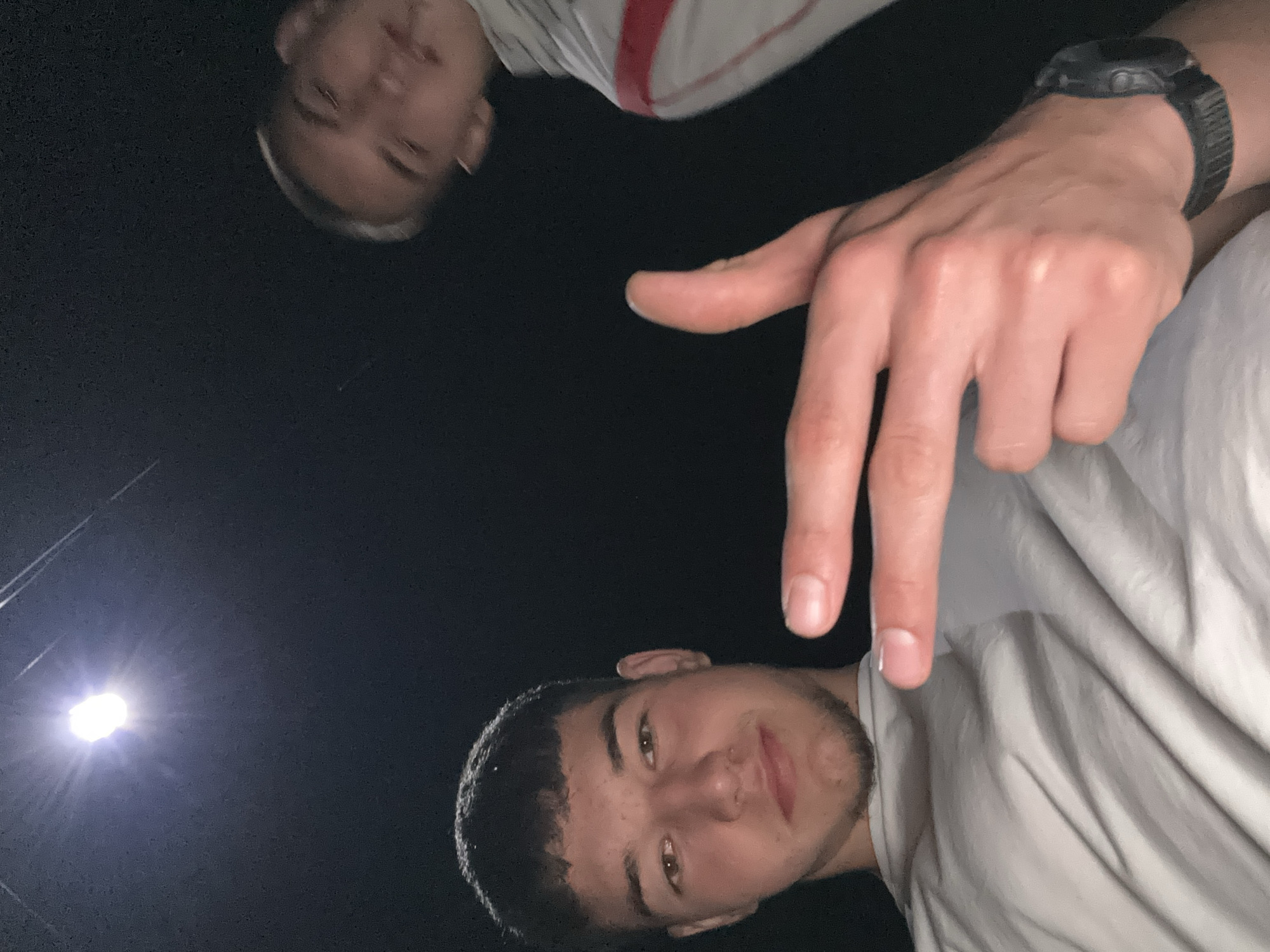 The image captures two young adult males at night in Zlatograd, Bulgaria, from a low-angle perspective. The primary subject, situated in the foreground, is a male with short, dark, textured hair, light stubble on his chin and jawline, and brown eyes. He wears a light-colored, possibly grey or white, short-sleeved t-shirt. His right hand is raised towards the camera, forming a gesture with his index finger and thumb extended, resembling a handgun. A dark digital wristwatch is visible on his left wrist. He looks directly at the camera with a slight smile. In the upper-right background, a second young adult male is partially visible and significantly out of focus. He wears a light-colored garment featuring a distinct red diagonal stripe and appears to be looking downwards or to the side. The scene is illuminated by a bright, artificial light source, likely an overhead street lamp, casting a strong glare in the upper-left corner of the frame. Faint, thin lines, possibly power or communication wires, are visible against the dark, featureless night sky.