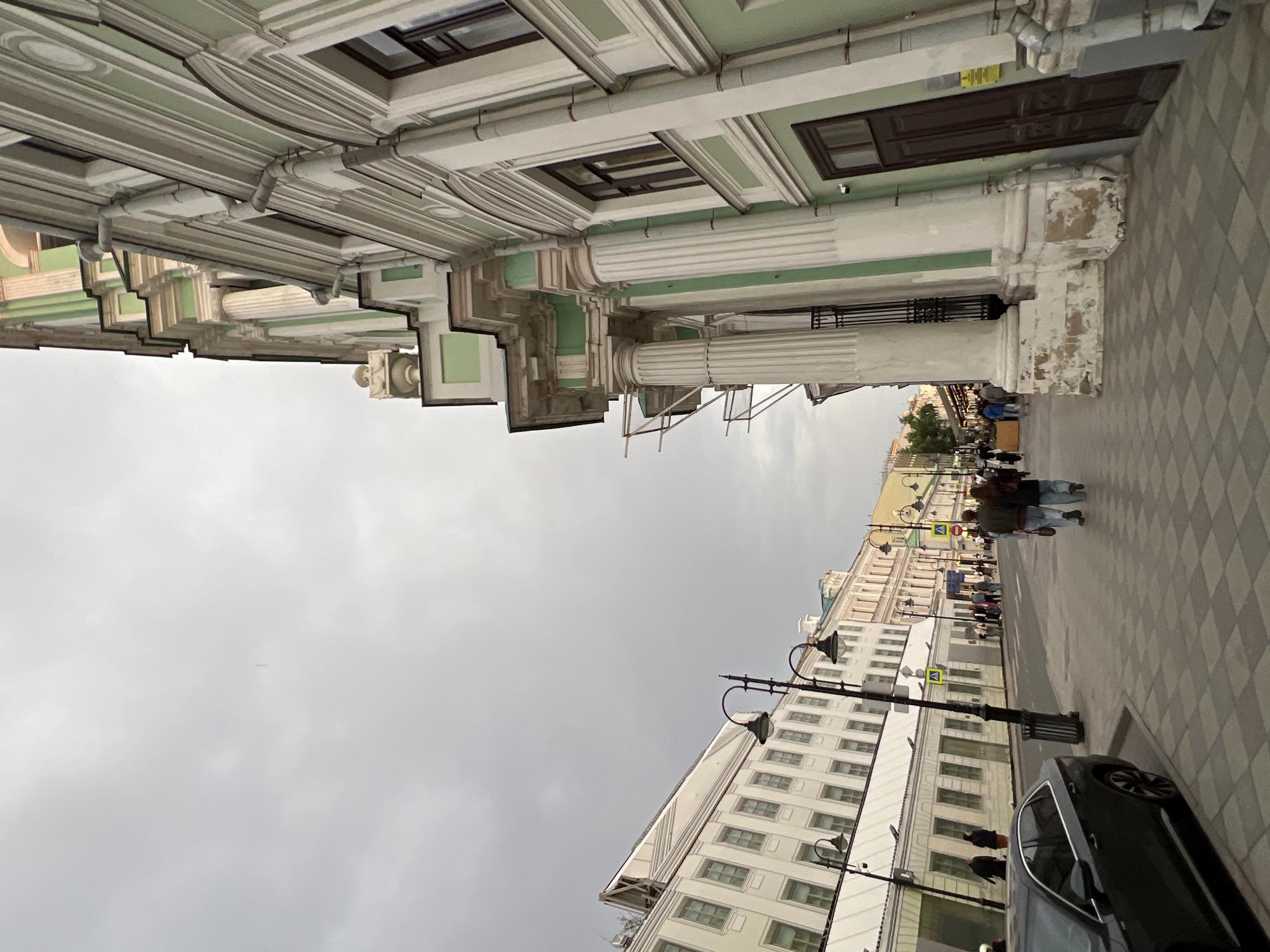 The image depicts a street scene in Moscow, Russia, characterized by classical architecture and overcast weather. On the right side, the facade of a building featuring decorative columns, ornate windows, and light green and white trim is visible. A sidewalk with a checkered pattern runs alongside the building, where several pedestrians are walking. On the left side of the image, another building with numerous windows and decorative street lamps is present. A portion of a dark-colored car is visible in the foreground. The sky is uniformly gray, indicating an overcast day.