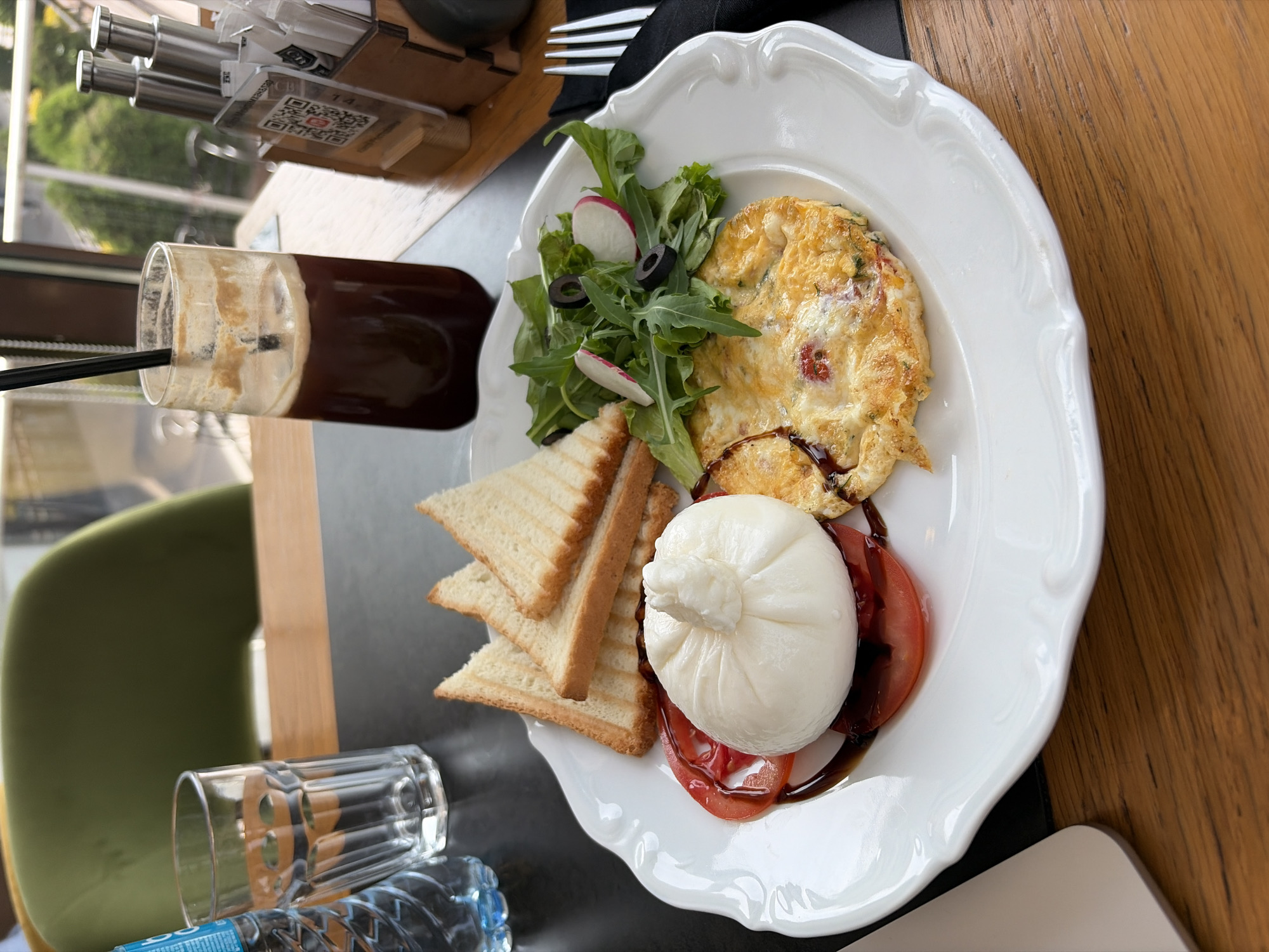 The image shows a breakfast meal served on a white ornate plate, placed on a table. The table appears to be located in a restaurant or cafe setting. The meal consists of burrata cheese on top of sliced tomatoes with a drizzle of balsamic reduction, a side of triangular toast slices, an omelet, and a small salad with radishes and olives. A beverage, possibly iced coffee, is visible in a tall glass with a straw. Cutlery is present near the beverage. In the background, there's a view of greenery through a window. The table also contains a bottle of water, an empty glass, and a container holding sugar or sweetener packets. Based on the background view of the outdoor environment and the style of the restaurant setting, this image might be taken in Almaty, Kazakhstan.
