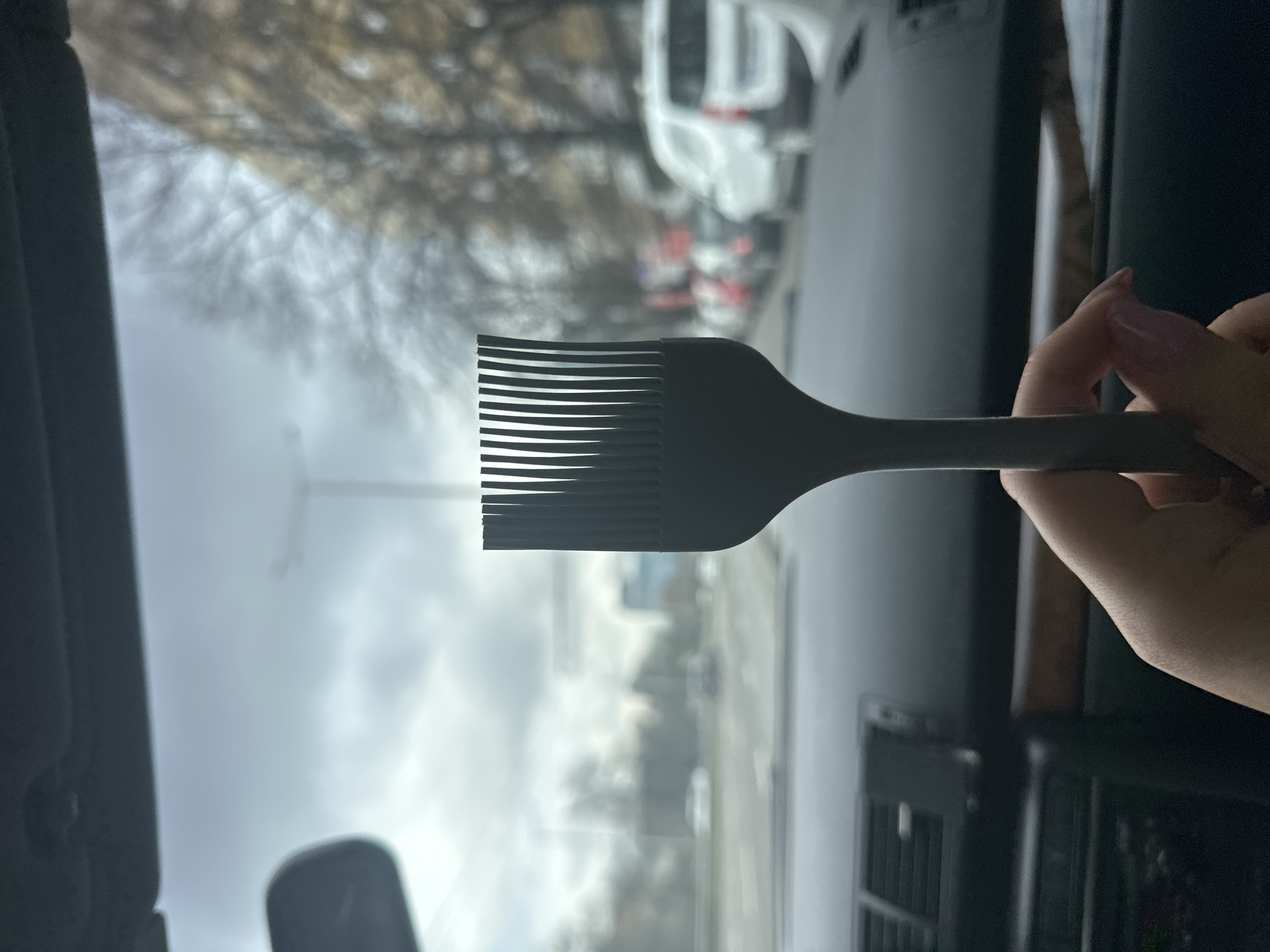 The image captures a close-up of a human hand, with light pink manicured fingernails, holding a dark grey, flat-bristled applicator brush vertically in the foreground. The scene is viewed from inside a vehicle, displaying a dark grey dashboard with visible air vents, a phone mount, and a wood trim accent. Through the vehicle's windshield, an urban streetscape in Sofia, Bulgaria, is visible under an overcast sky. The blurred background features a street with multiple lanes of traffic, including several cars and a white van. Bare deciduous trees line the thoroughfare, and indistinct multi-story buildings are discernible in the distance, along with a utility pole.