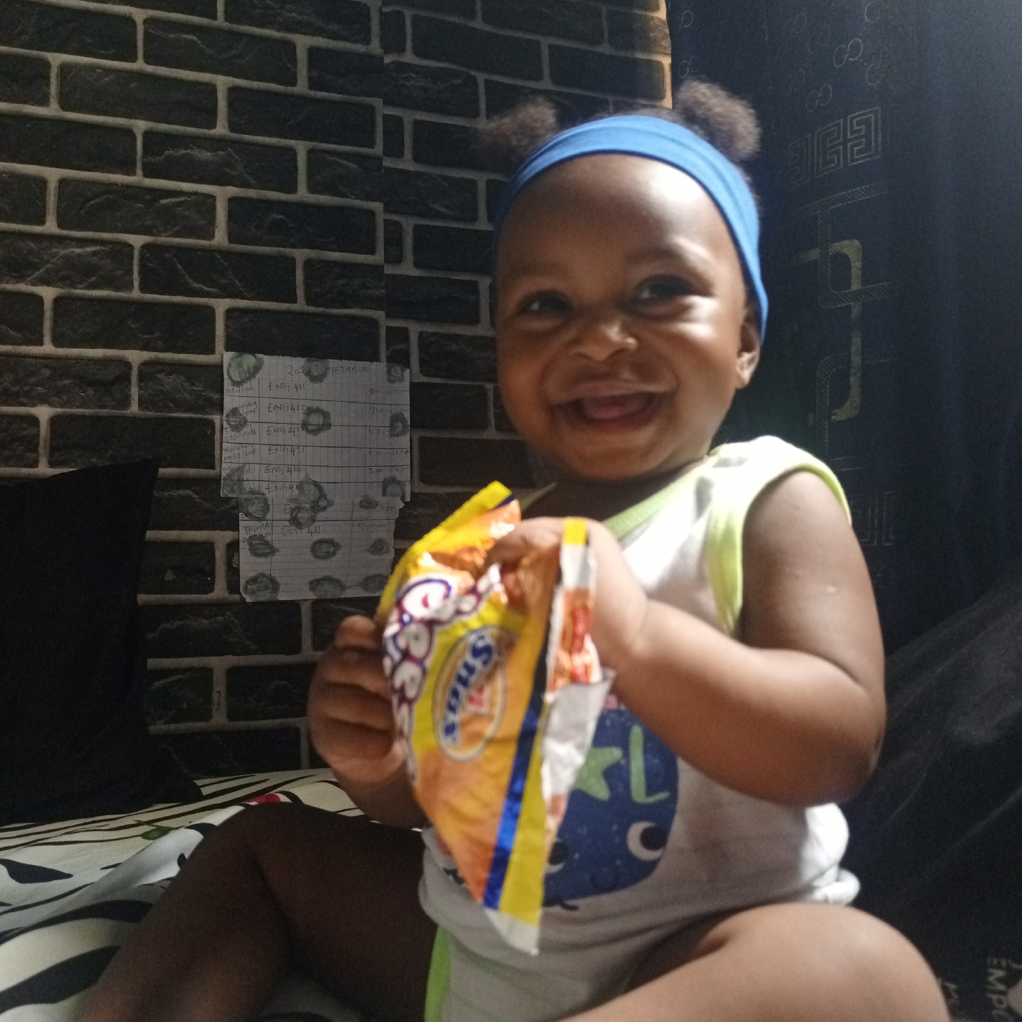 An indoor medium shot from Keffi, Nigeria, features a smiling baby or young child with dark skin and dark hair styled in two small puffs. The child wears a light blue headband and a white sleeveless top with a light green trim. A wide, joyful smile reveals an open mouth, conveying happiness. The child holds a yellow and orange snack bag, partially open, with the words "Sno" and "BeBe" visible.
The background on the left consists of a dark gray brick-patterned wallpaper. A piece of paper, likely an academic schedule or course list, is taped to the wall. Visible text on the paper includes "20 TETABL", "ENG 411", "ENG412", "09/09/2016 ENG 413", "10/11/05 ENG431", "ENG 414", "ENGAL", and "GST 411". To the right, a dark curtain or fabric hangs, displaying a repeating "G" or "GG" logo pattern, and the partial text "EMP" at the bottom. The child is seated on a bed covered with a white sheet adorned with black stripes and irregular patterns. A dark pillow or cushion is visible behind the child on the left. The lighting suggests an indoor setting, possibly dim, creating a cozy atmosphere.