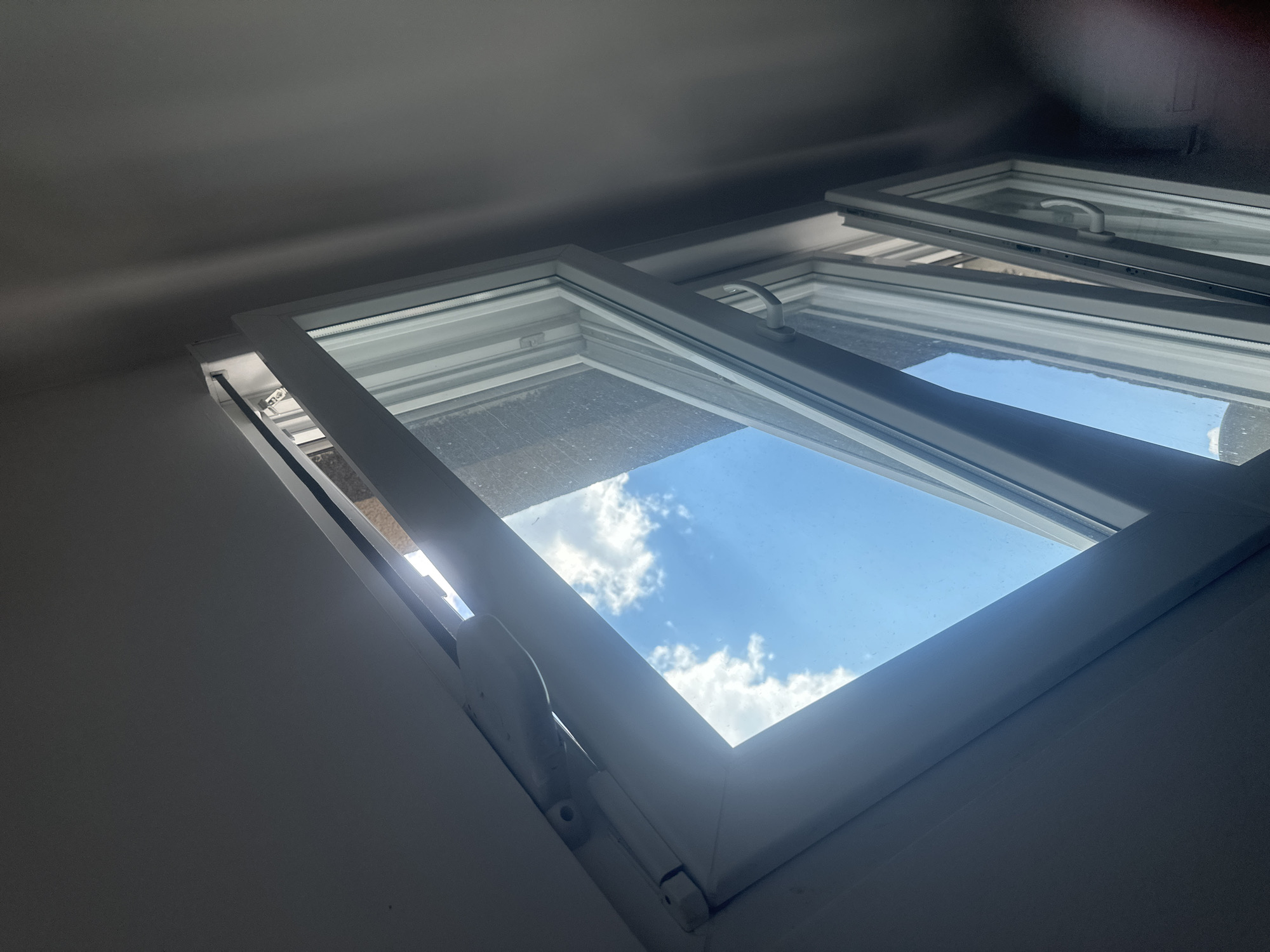 The image shows a view looking upwards through an open window. The window frame is white and appears to be made of uPVC. The window is partially open, with the top section tilted outwards. Through the window, a bright blue sky with fluffy white clouds is visible. The light suggests it is daytime. The interior of the room is dimly lit with a gray ceiling. No people are visible in the image. The scene is indoors looking outwards. The specific location in Zvezdara Urban Municipality, Serbia, cannot be determined from the visual information provided in the image.