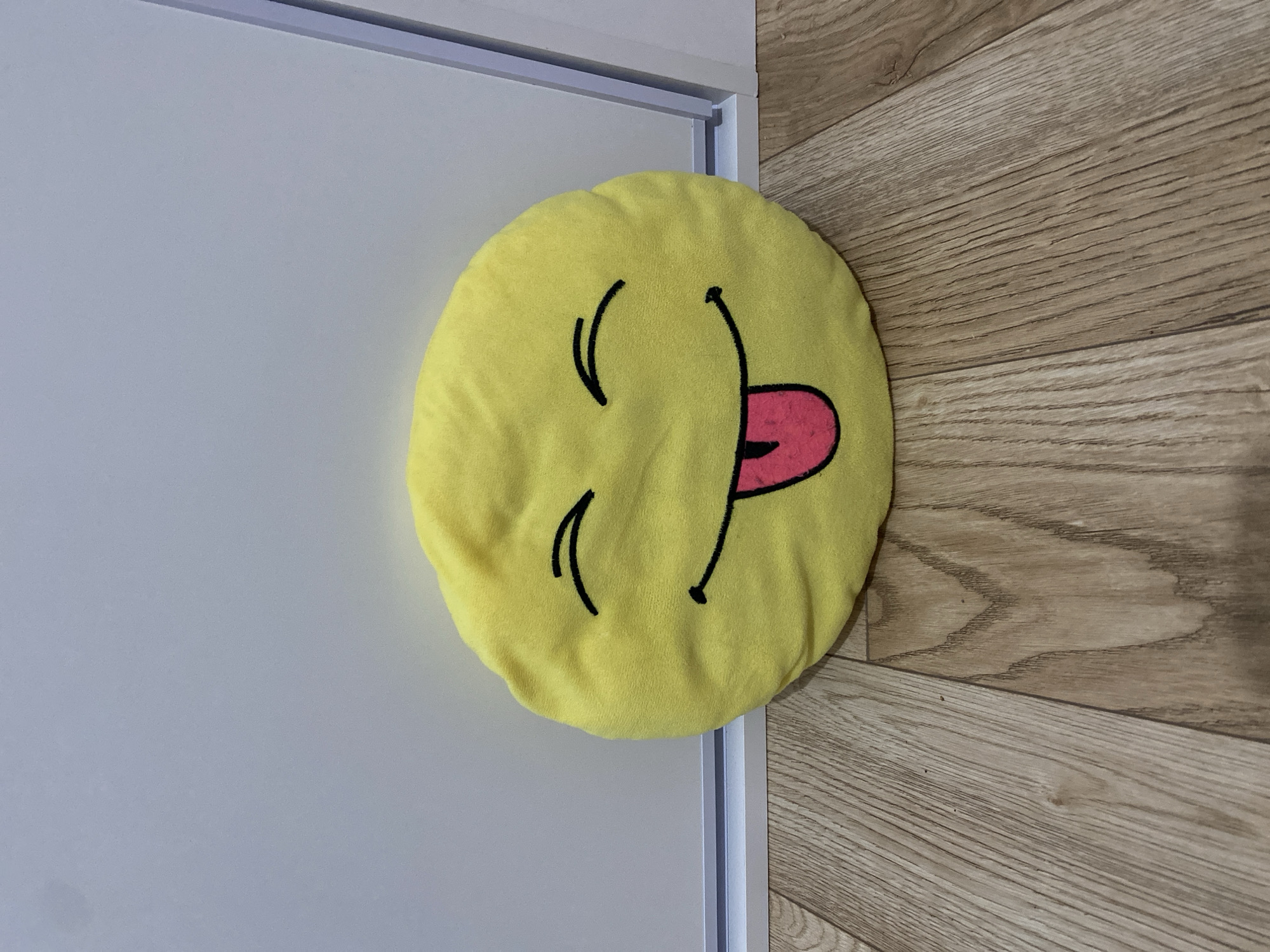 A yellow, plush, round pillow with a smiley face featuring closed eyes and a tongue sticking out is placed on a light brown wooden floor. The pillow is positioned in the lower center of the frame, against a plain, light gray wall. A white door frame is visible on the right side of the image, partially obscuring the wall. The lighting suggests an indoor environment. There are no people present in the image, and no specific details indicate the location is Minsk, Belarus.