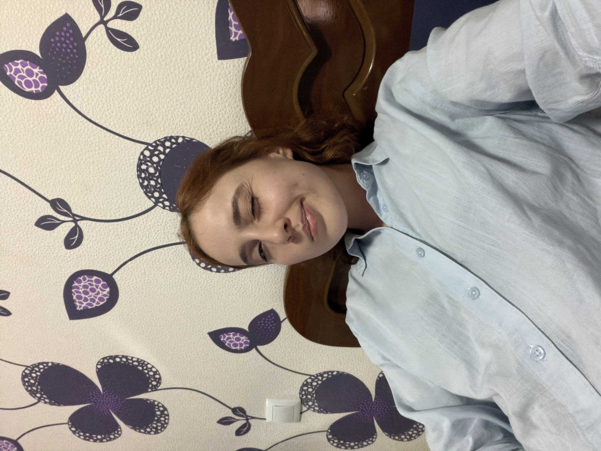 A young, light-skinned woman with auburn hair is lying in a bed or on a chair, wearing a light blue button-down shirt. She is smiling faintly with her eyes mostly closed, facing the viewer at an angle. Behind her is a wooden headboard or chair back. The wall behind the headboard is covered in a light beige wallpaper with a pattern of dark purple floral designs. There's an electrical outlet visible near the bottom of the frame on the wall. The setting appears to be indoors, likely a bedroom. There is no information to indicate the location in the city of Tbilisi, Georgia.