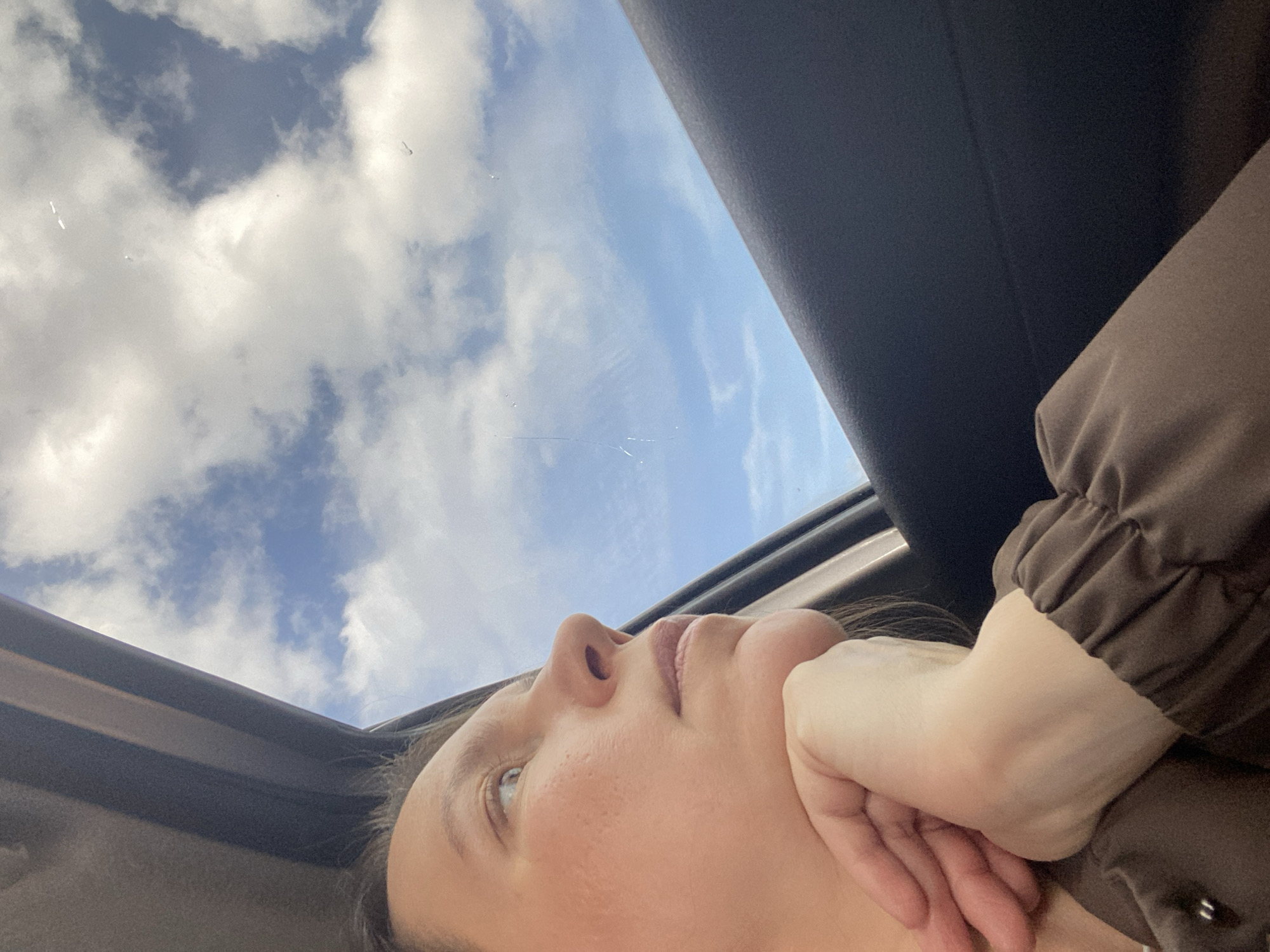 A low-angle shot shows a person lying in a car, gazing upwards through an open sunroof. The person's face is visible, featuring their nose, lips, and one eye which is looking towards the sky. Their hand is positioned under their chin, supporting their head. The sky is visible through the sunroof, displaying a bright blue hue interspersed with white, puffy clouds. Parts of the car's interior, including the sunroof frame and the roof lining, are also visible, appearing in dark shades. The person is wearing a brown jacket with elasticated cuffs. The location of Alnashi, Russia is not discernible from the provided visual information.