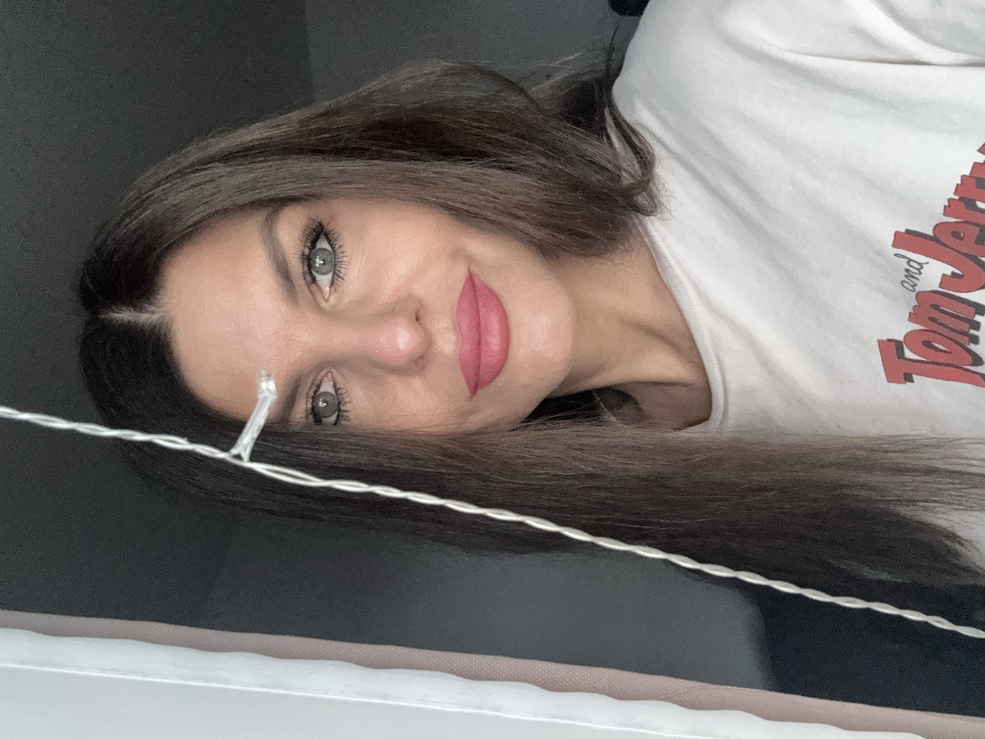 A woman with long, dark brown hair and light green eyes is looking at the camera. She is wearing a white t-shirt with "Tom and Jerry" printed on it in red letters. Her lips are painted a bright pink. She is standing in front of a dark background, with a white curtain on the left side of the image. A string of white Christmas lights is draped across the front of her face. There is no discernible information about the location of this image in Chaikovsky, Russia.