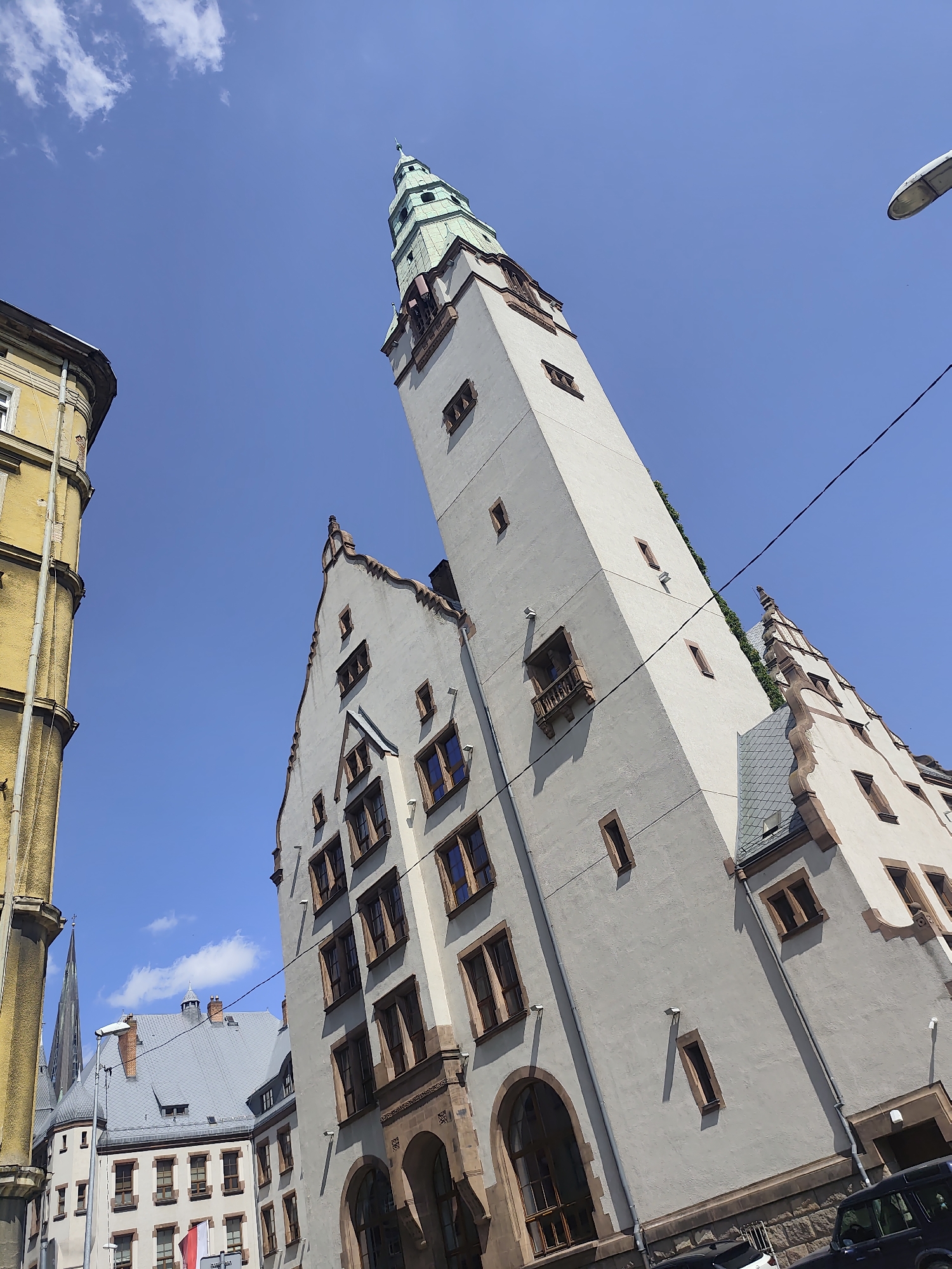 This is a low-angle, wide shot of the historic Collegium Minus building in Szczecin, Poland. The building's pale facade is dominant, featuring a tall, slender tower with a green, stepped spire. Rows of rectangular windows with dark frames are visible along the tower and the adjacent main building, which has a distinctive, stepped gable roofline. The building on the left, rendered in yellow, is partially visible and appears to be a separate structure, likely also historic. A street lamp is visible in the upper right corner, and power lines crisscross the bright blue sky, which has a few wispy clouds. The ground level of the Collegium Minus features arched entrances and is constructed with rough-hewn stone. The image suggests a bright, sunny day in an urban setting. There are no people clearly visible in the frame, and the overall impression is of an architectural study of the building.