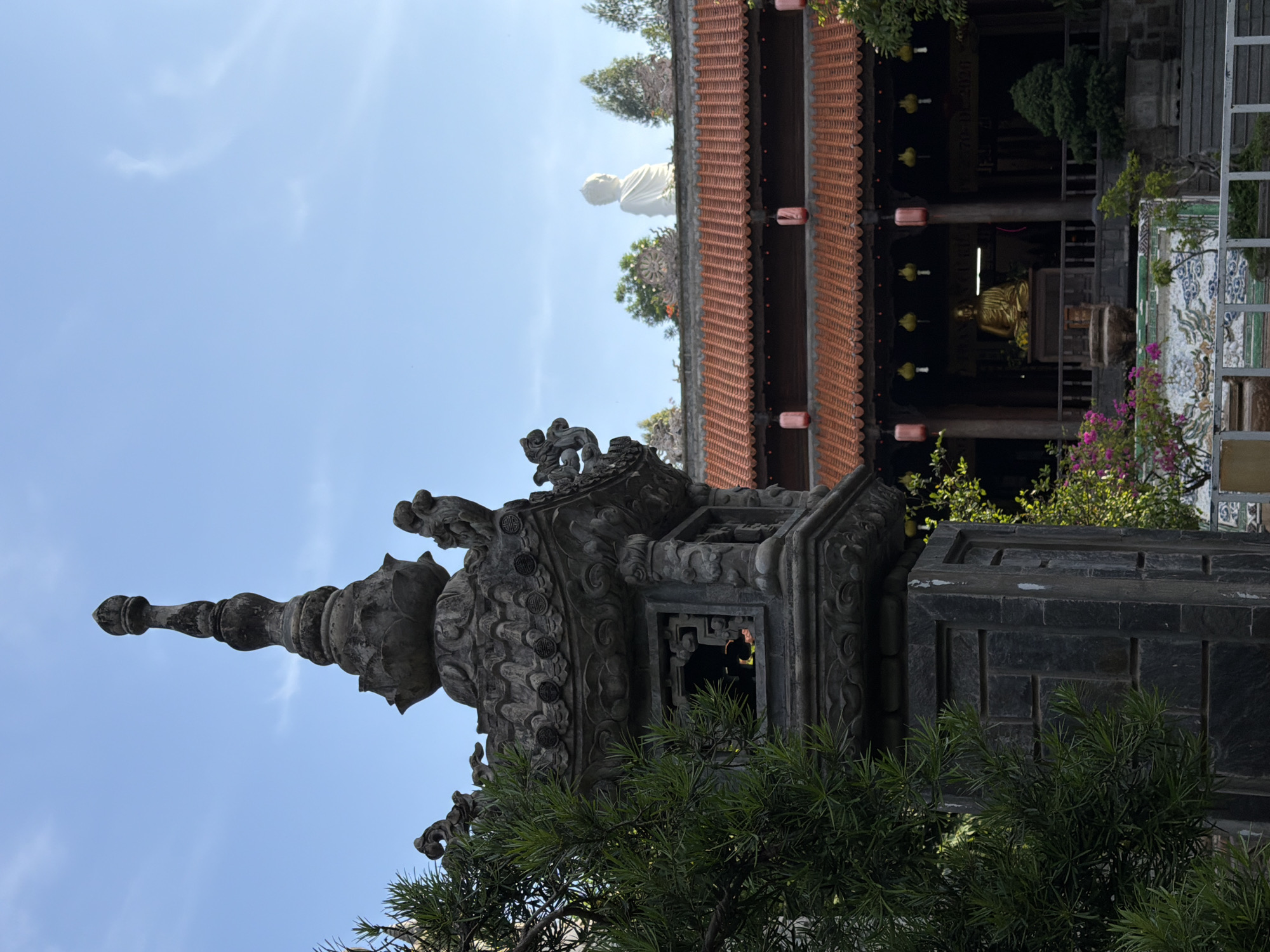 The image depicts a Buddhist temple in Nha Trang, Vietnam, under a clear blue sky. In the foreground, a weathered, ornate stone lantern stands to the left, partially obscured by lush green foliage. The lantern features intricate carvings and a hollowed-out section revealing a glimpse of yellow fabric within. Behind the lantern, the temple's architecture is visible, characterized by a red-tiled roof and traditional wooden structures.
Dominating the background is a large, white statue of Buddha situated on the temple's upper level, overlooking the scene. Below the white Buddha statue, a golden Buddha statue is seated within the main temple building, illuminated by ambient light. Red and yellow lanterns hang from the eaves of the temple, adding a festive touch. The temple grounds appear to be well-maintained, with potted plants and decorative mosaic work visible at the base of the building. The overall impression is one of serenity and cultural richness.