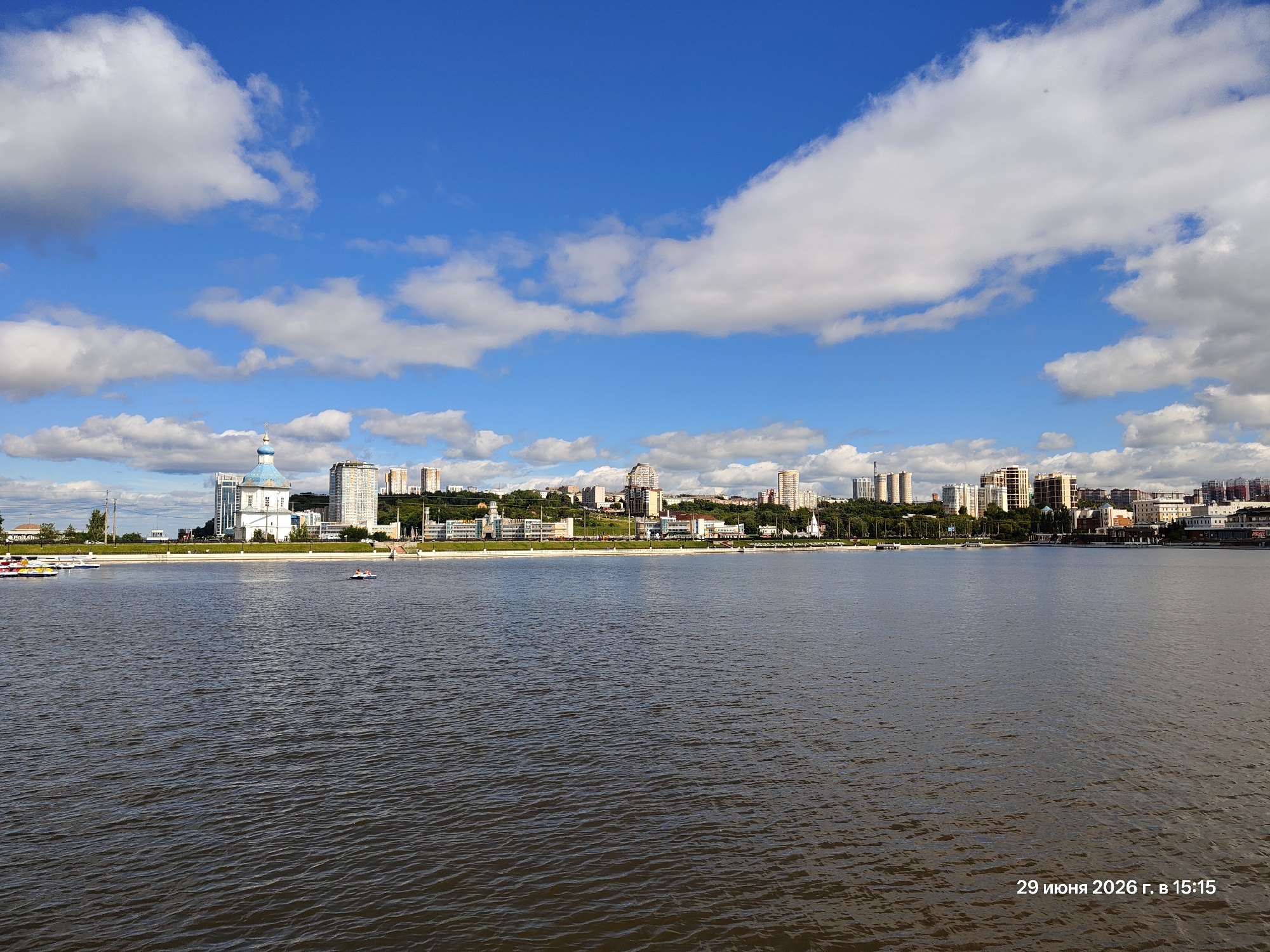 This is a photograph of a wide, calm river with a city skyline in the background. The water is dark and rippled, reflecting the blue sky and white clouds above. On the water, there are several small boats, including paddle boats docked on the left side of the image and a couple of small motorboats further out.
The city skyline is dominated by a mix of modern high-rise buildings and older, more traditional architecture. A prominent building with a blue dome and white walls stands out on the left side, appearing to be a church or other religious structure. Green trees and a grassy embankment separate the water from the urban landscape. The sky is a vibrant blue with scattered white clouds, suggesting a pleasant day.
In the lower right corner of the image, there is visible text in Russian. It reads: "29 июня 2026 г. в 15:15", which translates to "June 29, 2026 at 15:15". This indicates the date and time the photograph was taken. The overall mood of the image is peaceful and serene, capturing a moment of calm on the water with a city vista.