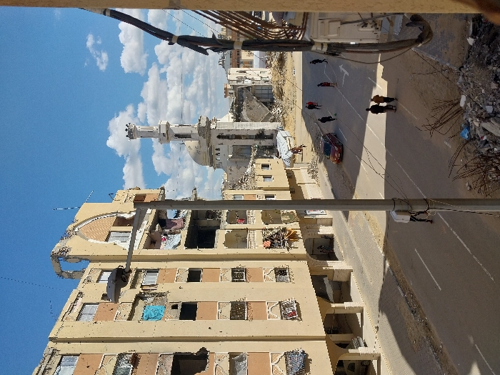 This image presents a daytime urban street scene in Khan Yunis, Palestinian Territory, under a blue sky with scattered white cumulus clouds.
On the left, a multi-story, light-colored residential building exhibits significant damage, characterized by large structural breaches, exposed interior elements, and missing facade sections. Some windows are open or boarded, and laundry is visible hanging from several openings. The ground floor features an arched colonnade.
In the center foreground, a grey utility pole with a street light rises, carrying numerous wires and casting a long shadow across the asphalt road. The road displays white lane markings, including an arrow. A reddish-brown SUV is present on the street, facing away from the viewer.
Approximately a dozen individuals are visible: two adults walk together in the mid-ground, a group of five adults is further down the street, one individual is near the base of the utility pole, another individual walks towards the damaged building on the left, and a small group is discernible under the arched colonnade of that building.
In the background, a white mosque with a prominent dome and a tall, slender minaret stands. The mosque shows structural damage, with sections of its facade missing. Surrounding and beyond the mosque are extensive areas of rubble and partially destroyed buildings, indicating widespread urban devastation. In the lower right foreground, a pile of concrete debris and twisted rebar is present. The scene is framed on the right by a vertical building element with attached cables.