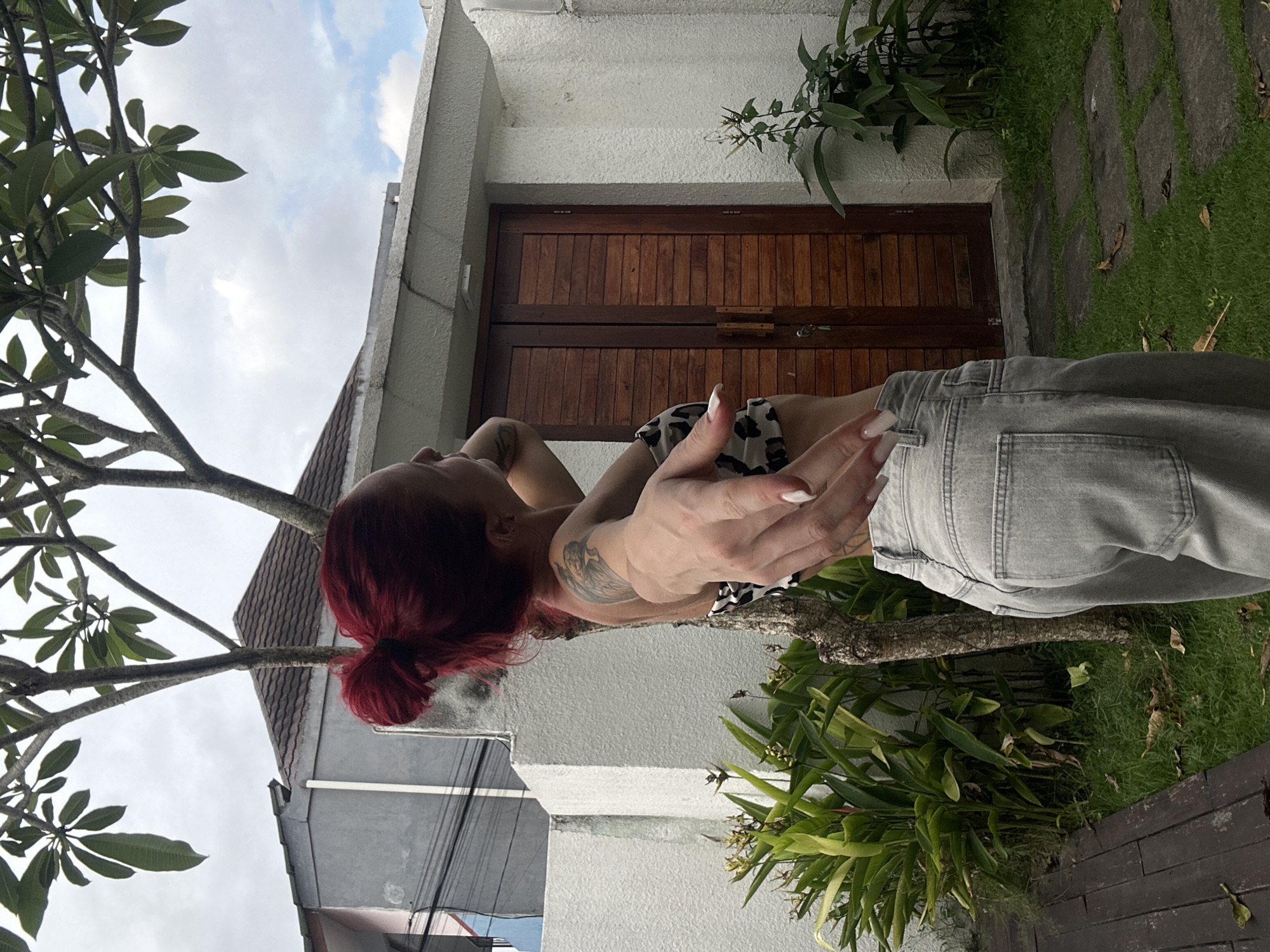 The image, taken in Dalung, Indonesia, shows a woman with red hair in a bun and a tattoo on her left arm. She is wearing a spotted top and light grey jeans. She is positioned in front of a white building with a brown wooden door and a tiled roof. A tree with green leaves is visible on the left side of the frame, and greenery is present around the building. The ground is covered with grass and stone paths.