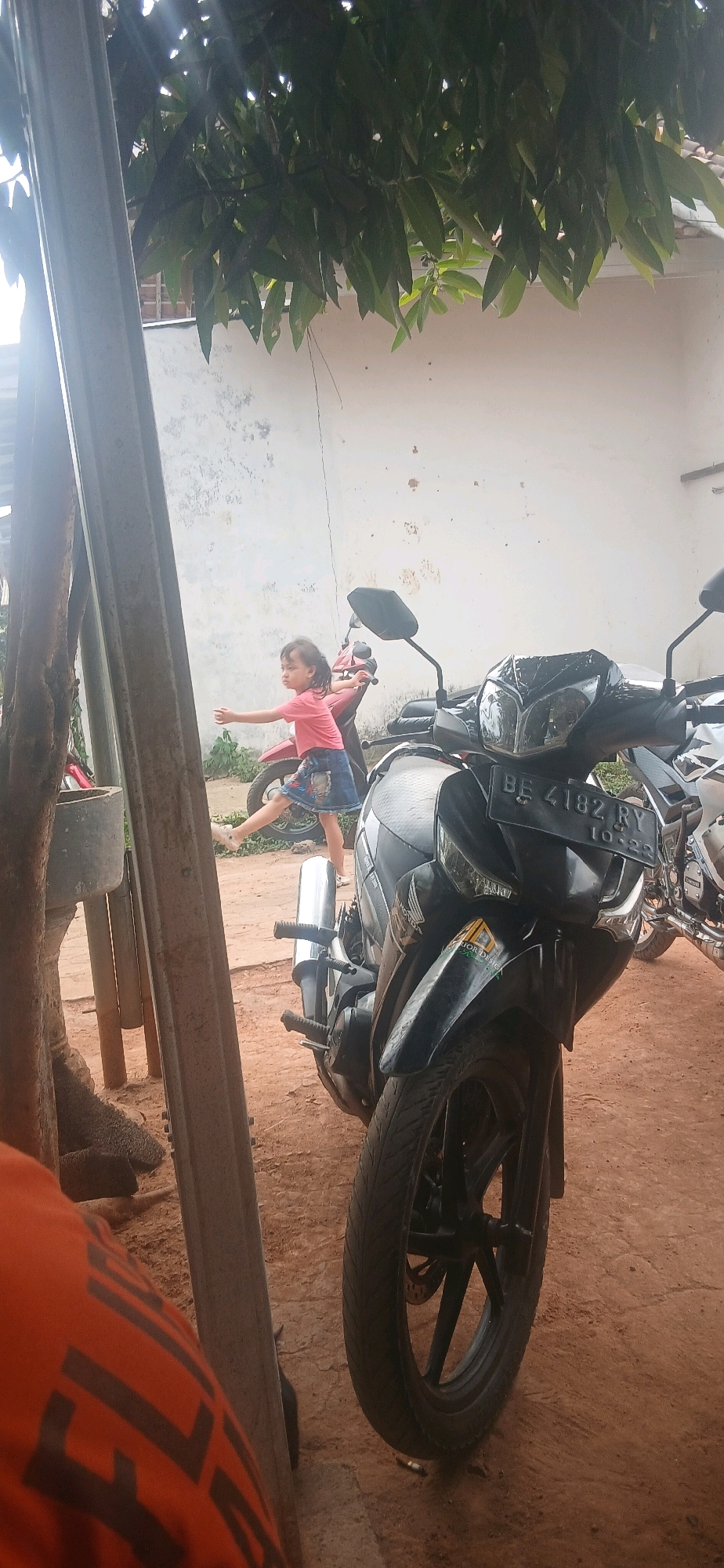 A low-angle, outdoor, eye-level shot shows a young girl in a pink shirt and denim skirt mid-stride, her arms outstretched to her sides, as if about to leap. She is standing next to a red motorcycle, with a black motorcycle in the foreground, obscuring the lower portion of the image. A tall, weathered metal pole runs along the left side of the frame, and large leaves from a tree canopy cast dappled shadows across the scene. The background consists of a textured white wall and a dirt ground. The license plate on the black motorcycle reads "BE 4182 RY." The image appears to have been taken in a residential area in Metro, Indonesia.