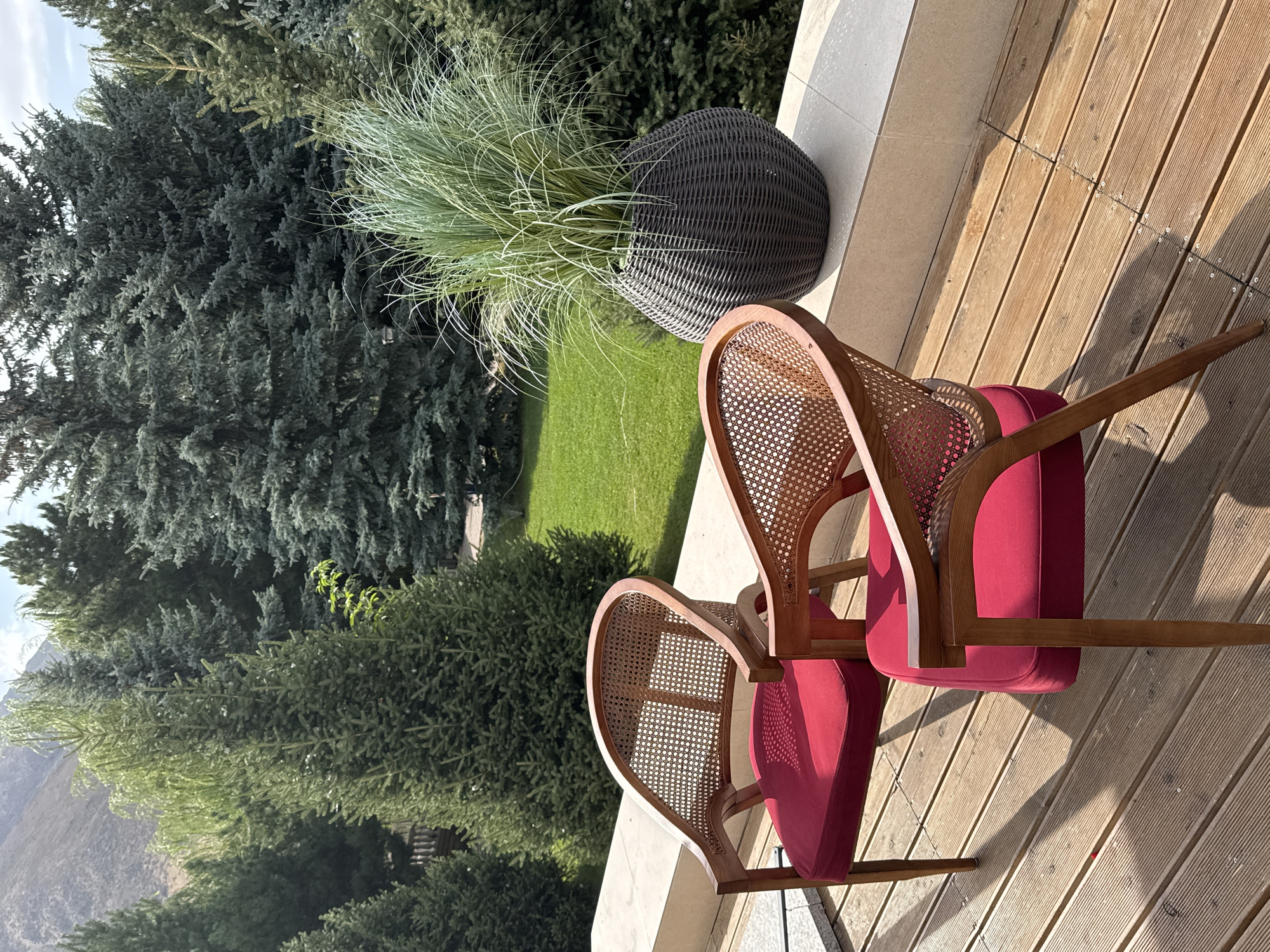 The image depicts a serene outdoor scene with two elegant cane-backed chairs featuring vibrant red cushions, positioned on a wooden deck. In the background, a lush green lawn is bordered by a dense forest of evergreen trees, with a glimpse of a mountain range in the distance. A large, dark woven planter filled with feathery green grass sits on a beige stone ledge, completing the tranquil ambiance. The location is identified as Koy Tash, Kyrgyzstan.