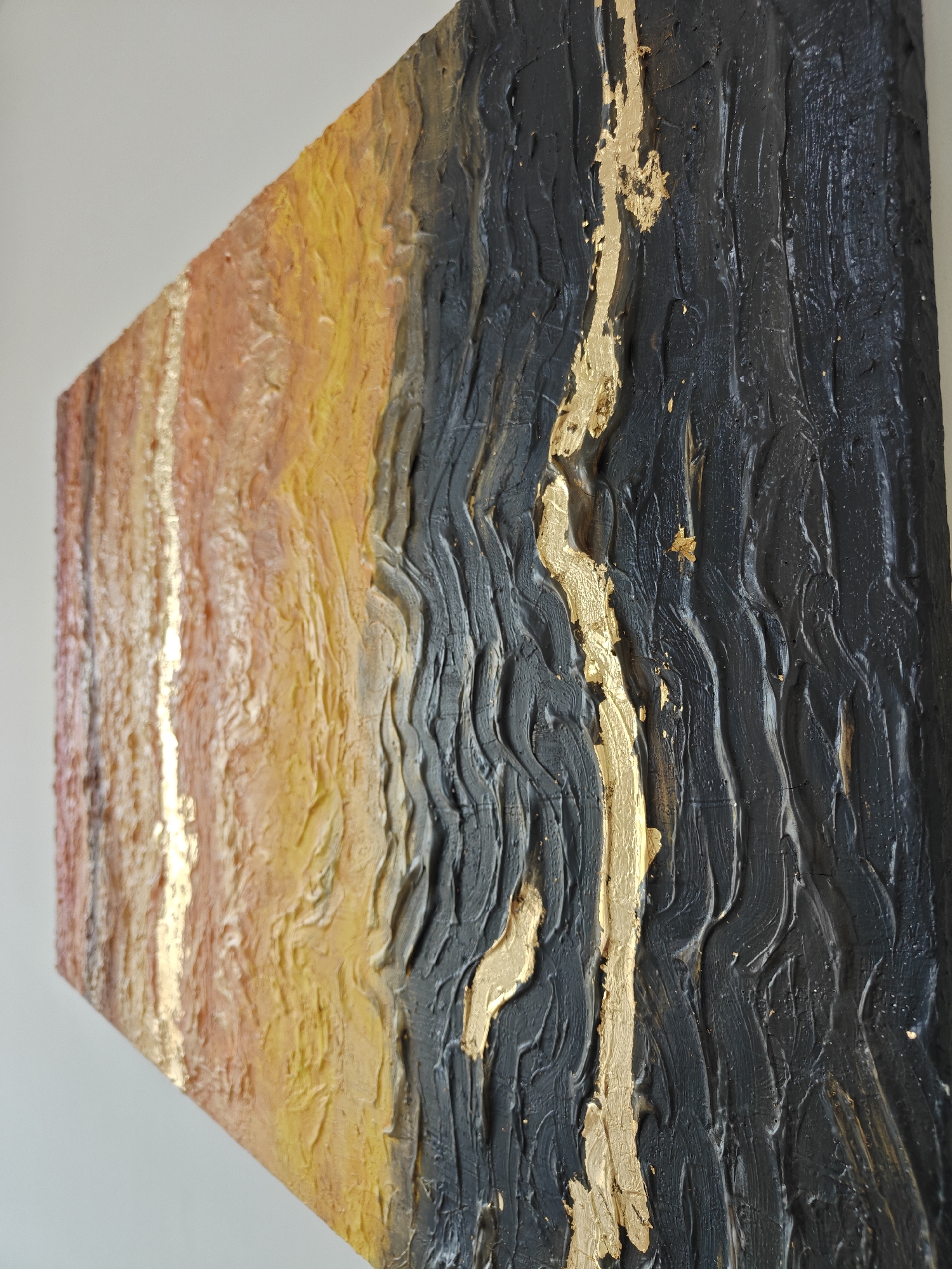The image is a close-up of a textured, abstract painting. The painting has a distinct color gradient, transitioning from shades of red and yellow on the left side to dark gray and black on the right. A streak of gold leaf is applied vertically across the painting. The texture is created by thick layers of paint, forming ridges and valleys that run parallel to the color gradient. The photo is taken at an angle, showcasing the depth of the painting. The location in Mumbai, India is not evident in the image.
