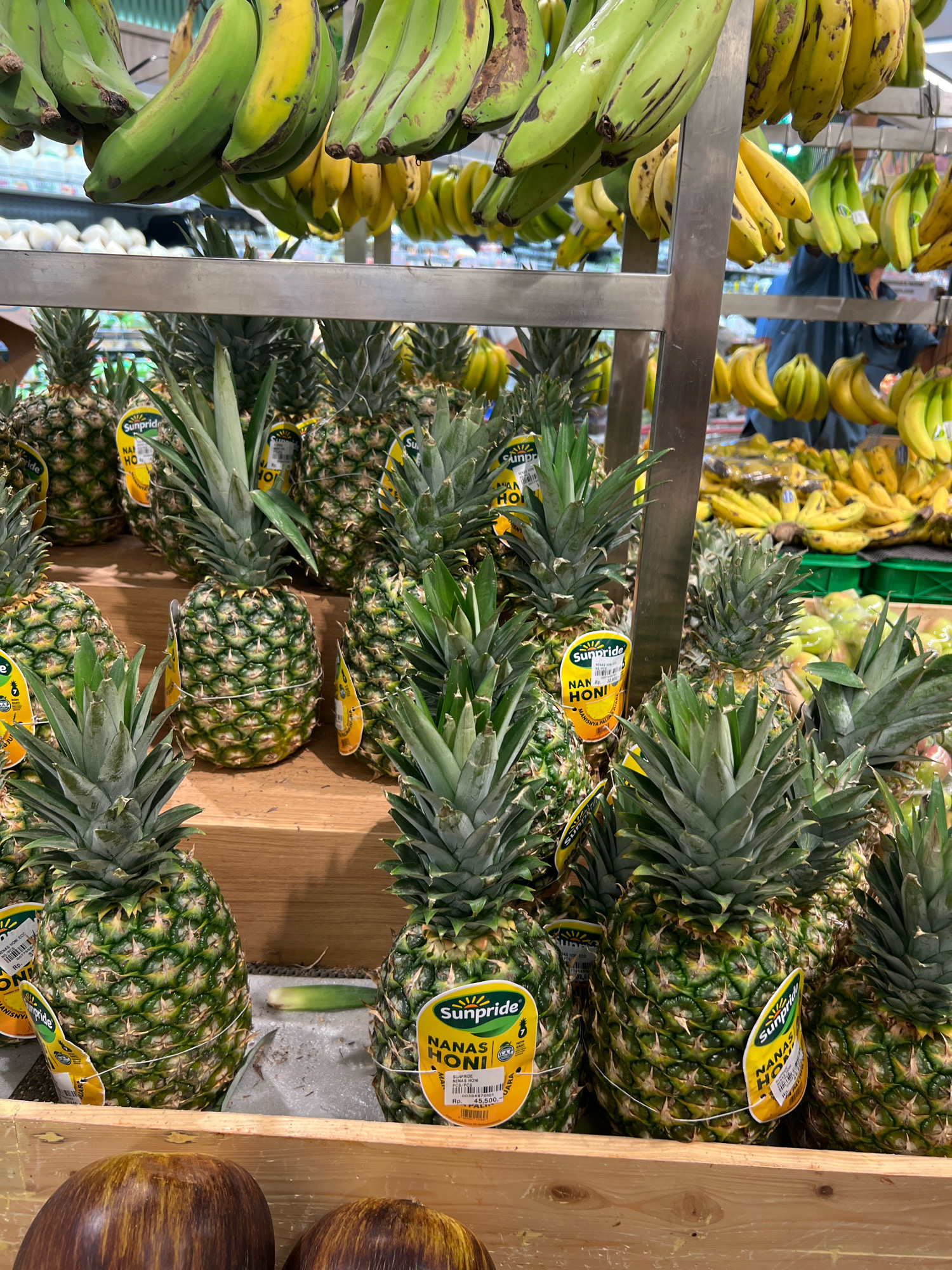 The image shows a produce display at a market or grocery store. In the foreground, there are several large pineapples stacked in wooden crates. Some of the pineapples have yellow labels with the brand name "Sunpride" and text in Indonesian, including "NANAS HONI" (Honey Pineapple). Behind the pineapples, there are bunches of green and yellow bananas hanging from a metal rack. Further in the background, more bananas and some green fruits, possibly apples, are visible in bins. A person wearing a blue shirt is partially visible in the background. The overall environment appears to be an indoor market with shelves and displays of fresh produce. Based on the presence of "NANAS HONI" labels, which indicates a product likely sold in Indonesia, and the general appearance of the market, this scene could be in Kuta, Indonesia, a popular tourist destination known for its markets.