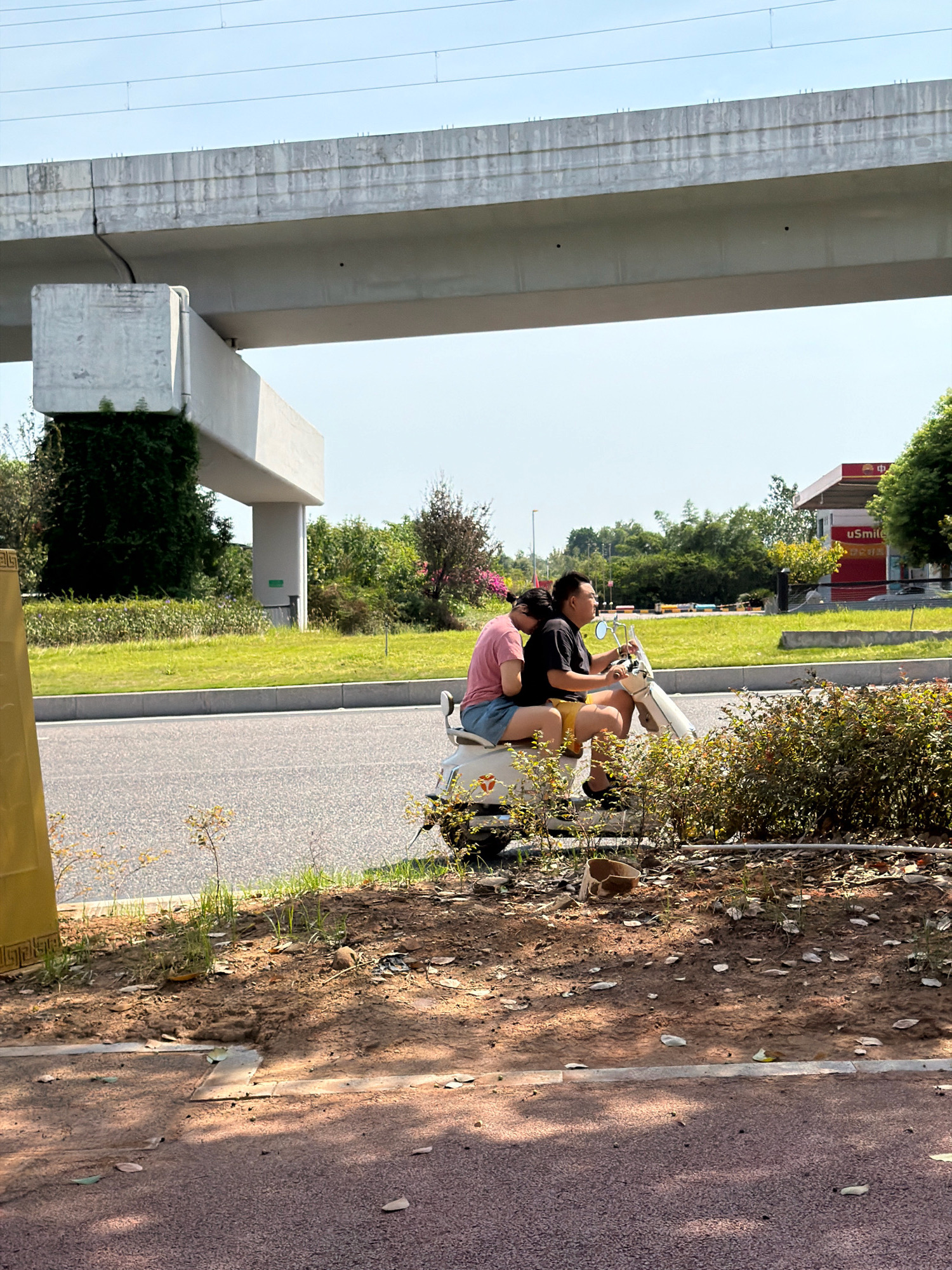 In Jiangjin, China, two individuals are riding a white scooter on a paved road under a concrete overpass. The person in the back, a woman with dark hair pulled back, is leaning on the shoulder of the person in the front, a man with dark hair. She is wearing a pink t-shirt and blue shorts. He is wearing a black t-shirt and yellow shorts. The scooter has a white body and a brown seat. To the right of the scooter, there are green bushes. In the background, there is a red and white gas station with a sign that says "uSmile". The sky is clear and blue. The overall scene is a snapshot of daily life in an urban environment.