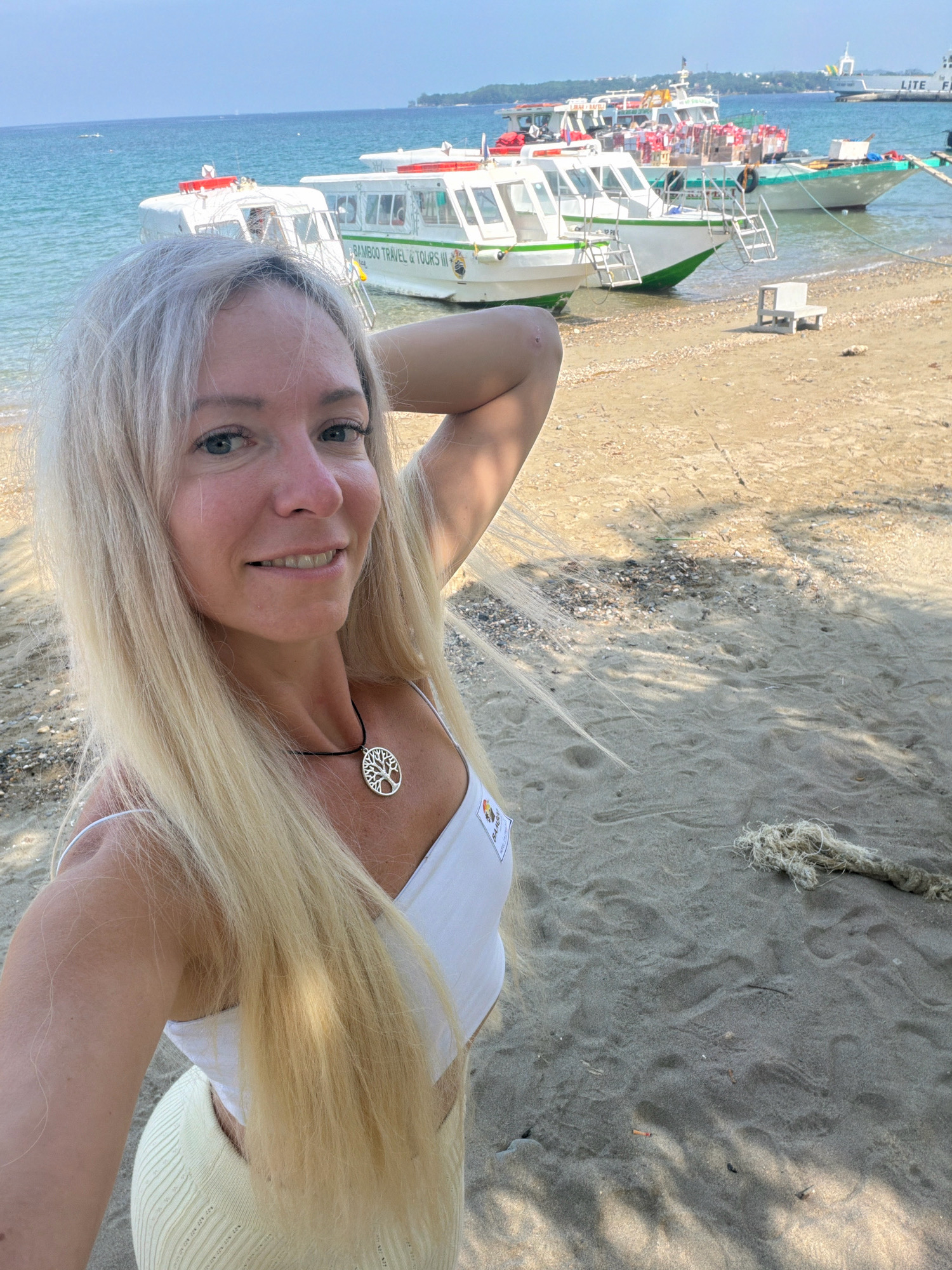 In this full-frame, outdoor, daytime photograph taken in Malay, Philippines, a fair-skinned Caucasian woman with long, blonde hair smiles into the camera. She is wearing a white tank top and a light yellow ribbed skirt. A silver tree of life pendant hangs from a black cord around her neck. Her left arm is raised and bent, with her hand resting on the back of her head.
The background features a sandy beach with a few scattered shells and debris. Several white passenger boats are docked in the shallow, blue-green water. The boats have green hulls and white canopies. One boat has the words "BAMBOO TRAVEL & TOURS III" printed on its side. A small, white plastic bench sits on the sand to the right of the boats. In the distance, the coastline with some vegetation and buildings is visible, and the sky is clear and blue. The lighting suggests a sunny day, with shadows from unseen trees falling across the sand.