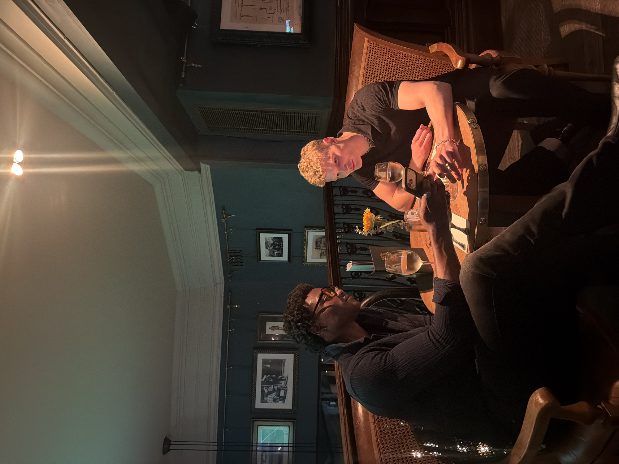 The image depicts two men seated at a wooden table in a dimly lit, atmospheric interior, likely a restaurant, bar, or pub in London, during the evening. The setting suggests a warm and intimate ambiance.
One man, with light, curly hair, wearing a dark t-shirt and a silver chain, is positioned on the left side of the table. He is looking towards the other individual, with his hands resting near a smartphone and a glass of white wine. The second man, with dark, curly hair and glasses, wears a dark, textured long-sleeved shirt. He is holding a smartphone and appears to be looking upwards or engaging in conversation with the first man, with another glass of white wine nearby.
A small vase containing a sunflower is placed on the table between them. The wooden table features metallic stud detailing along its edge. The chairs have classic designs with woven cane backs. The dark-colored walls are adorned with various framed pictures, and architectural details like ceiling moldings and a ventilation grate are visible. Warm artificial lighting casts highlights on the subjects, drinks, and table, with lens flares visible from light sources on the left side of the frame. A small tag or card displaying "04" is visible near one of the wine glasses. The overall scene suggests a relaxed social gathering.