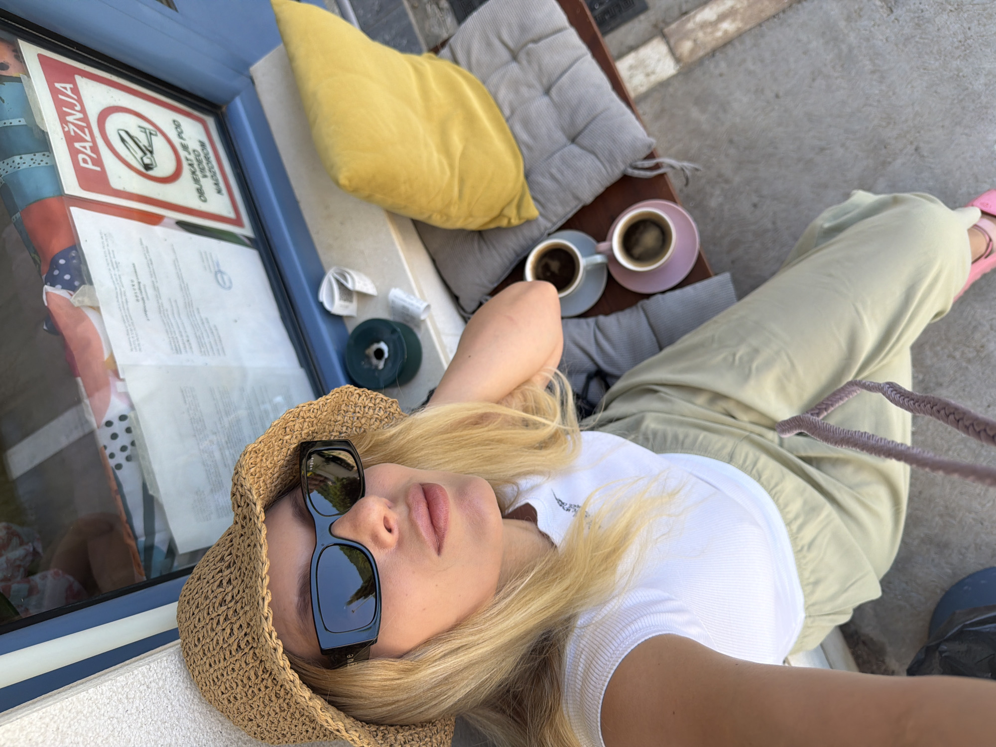 The image shows a woman lying on a bench outdoors, with a straw hat and sunglasses on. She is wearing a white t-shirt and light-colored pants. There are two cups of coffee on saucers, a yellow pillow, and a grey cushion on the bench. A sign with red and white lettering is visible to the left, reading "PAŽNJA" and "OBJEKAT JE POD VIDEO NADZOROM." This translates to "ATTENTION" and "THE OBJECT IS UNDER VIDEO SURVEILLANCE." The scene appears to be in Budva, Montenegro, judging by the visible text and the general aesthetic. The lighting suggests it is daytime, likely sunny. The woman's pose and the presence of coffee imply a relaxed moment, possibly a break or leisure time. The background shows a glimpse of an outdoor paved area and some greenery.