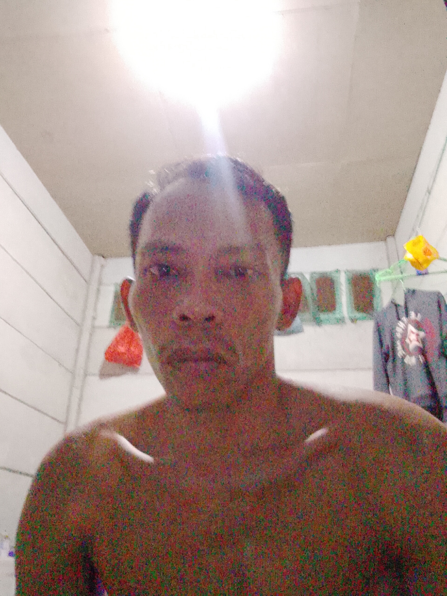 A man is taking a selfie in a room in Bukit Ketok, Indonesia. He is shirtless. Behind him is a white wall with what appears to be clothing hanging on a clothesline. The room has a white ceiling with a bright light source above. The image is slightly out of focus with visible digital noise.