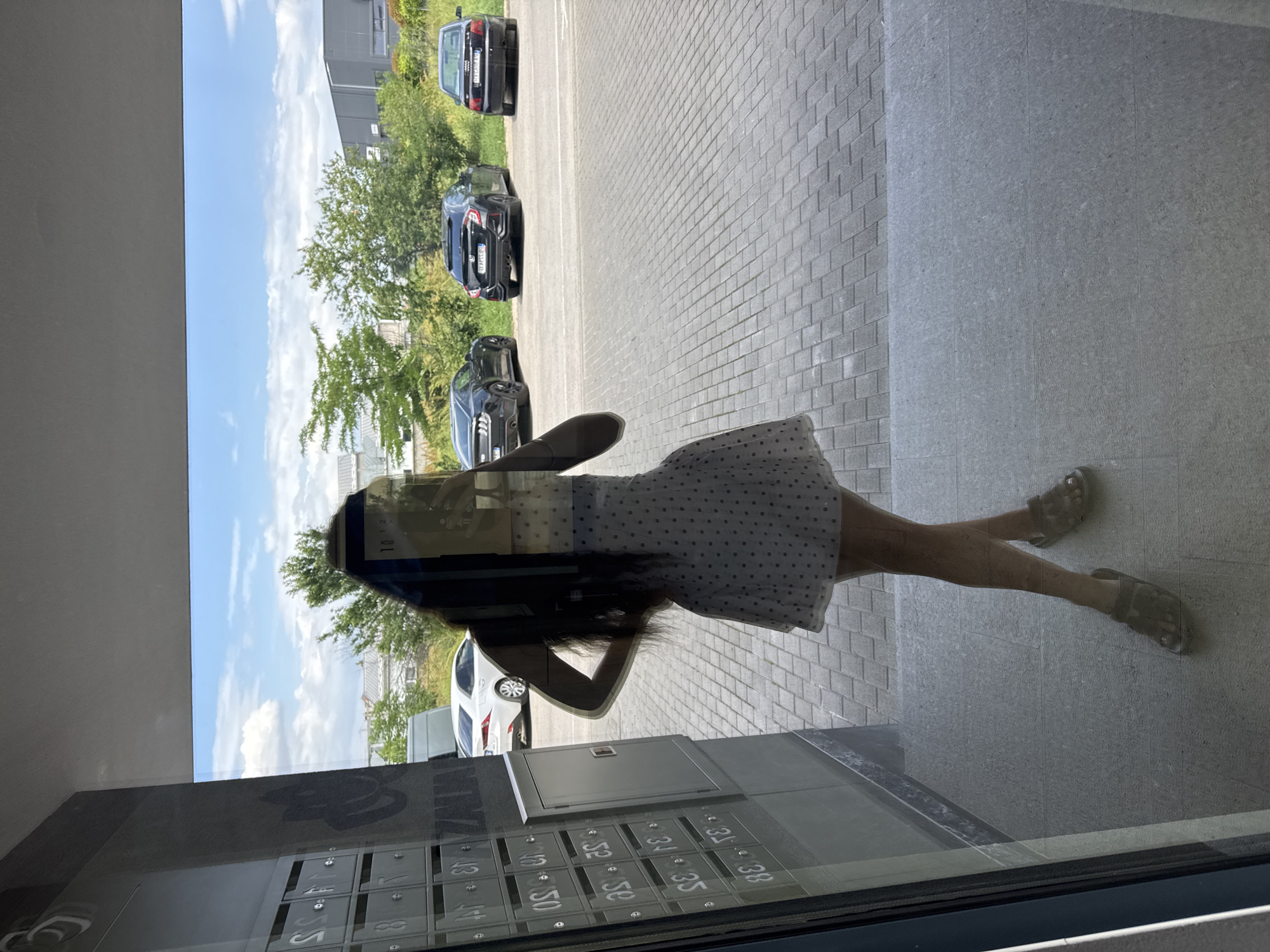 A reflection in a window shows a woman in a white dress with dark spots and sandals standing on gray tile. The reflection also shows a row of mailboxes below a gray wall, a paved area outside, a street with cars, trees, and a building. The location is Plovdiv, Bulgaria.