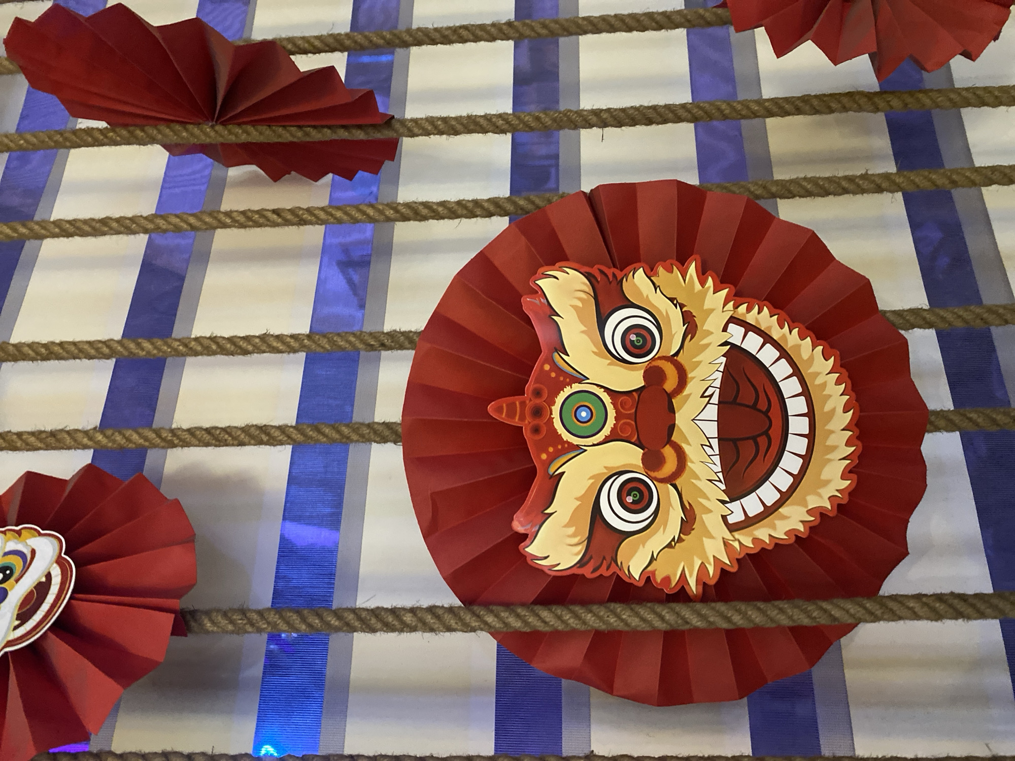 The image shows a decorative display of red paper fans, some featuring lion faces, hanging vertically against a backdrop of blue and white stripes. Thick, braided ropes are interspersed among the decorations, also hanging vertically. The overall impression is festive and possibly related to a cultural celebration. The image's metadata does not contain information about its specific location in the city of Đà Nẵng, Vietnam.