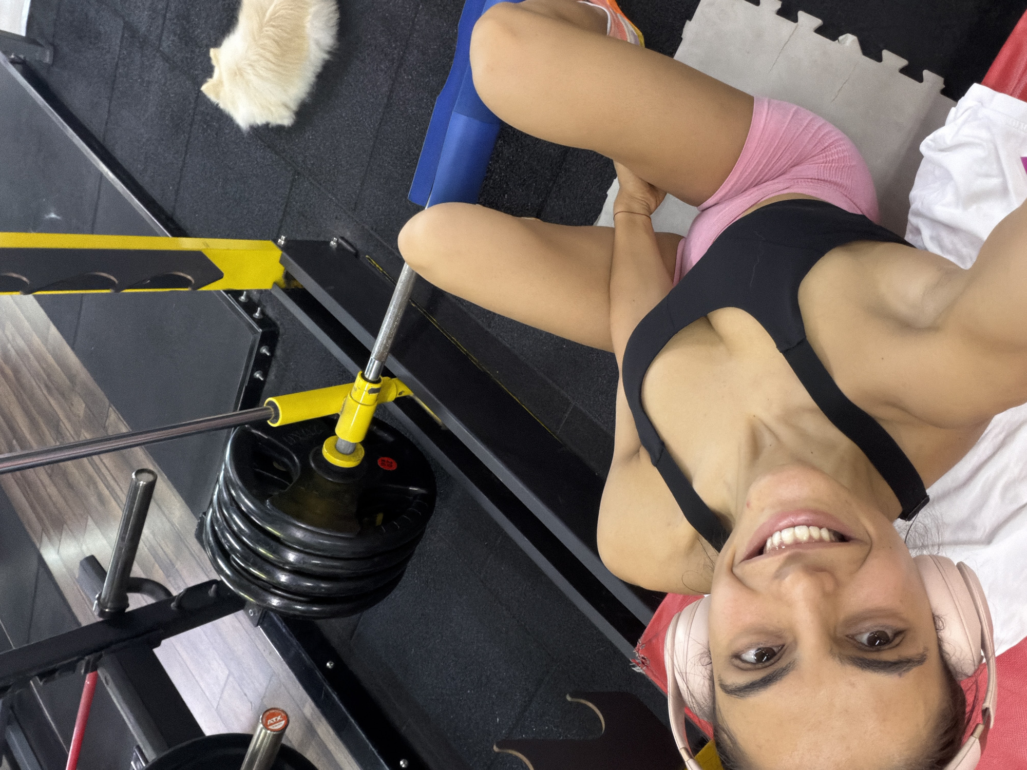 A woman with dark hair and brown eyes, wearing pink headphones and a black sports bra with pink shorts, lies on her back on a white mat on the floor of a gym. She is smiling at the camera. Behind her, a barbell with weight plates is loaded onto a weight machine. A small, fluffy light-colored dog is lying on the black rubber floor to the right of the weight machine. The gym floor is black rubber, and the walls are a neutral color. The lighting is bright and even. The location is Svilengrad, Bulgaria.