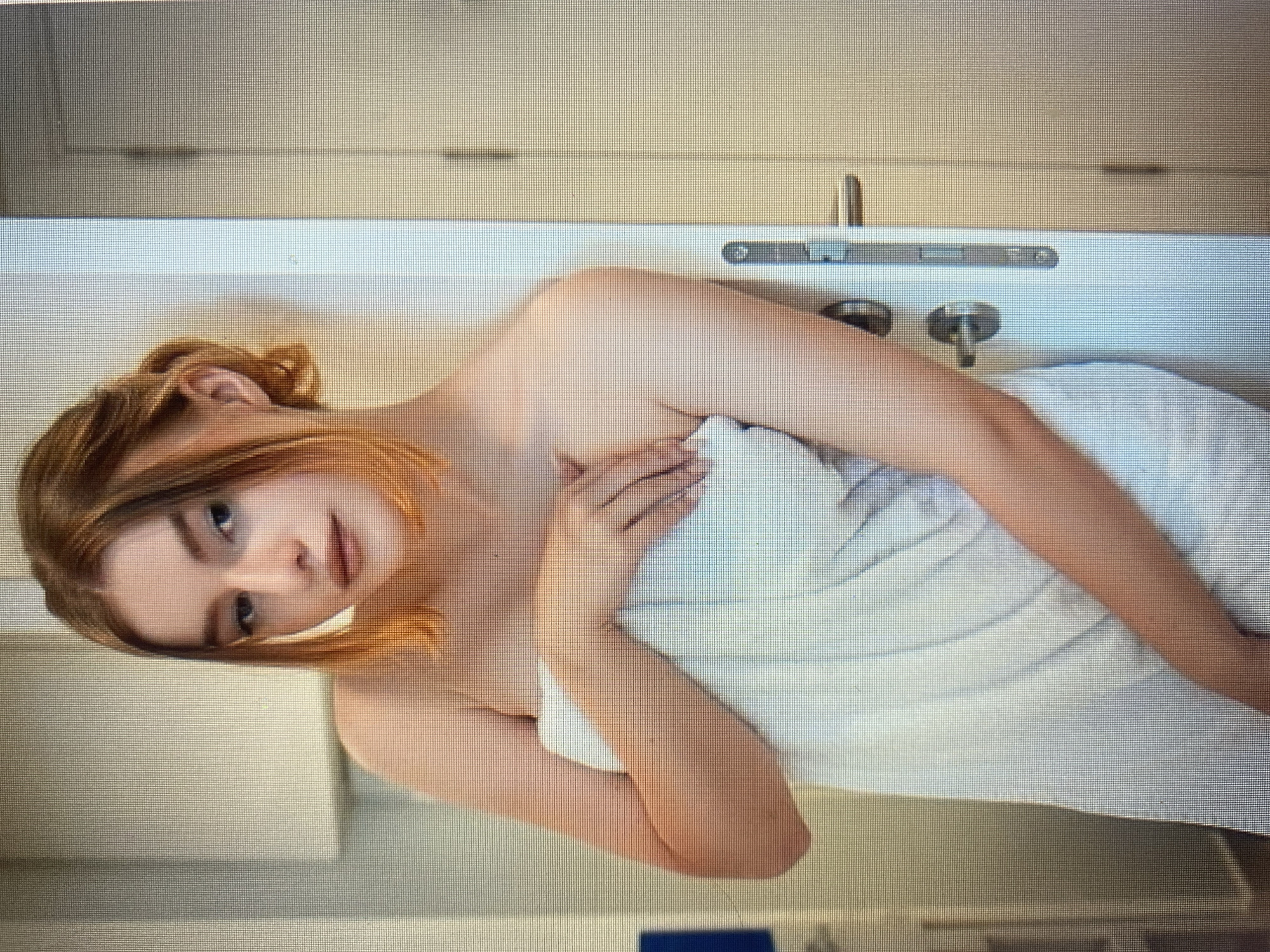 A young woman with auburn hair, wearing a white towel, leans against a white doorframe. Her hair is pulled back loosely, with some strands framing her face. She is looking towards the camera with a soft expression. Her right arm is bent, with her hand resting on the towel near her chest. The doorframe features a silver door handle and lock. The background appears to be an indoor setting with a blurred light-colored wall on the left.