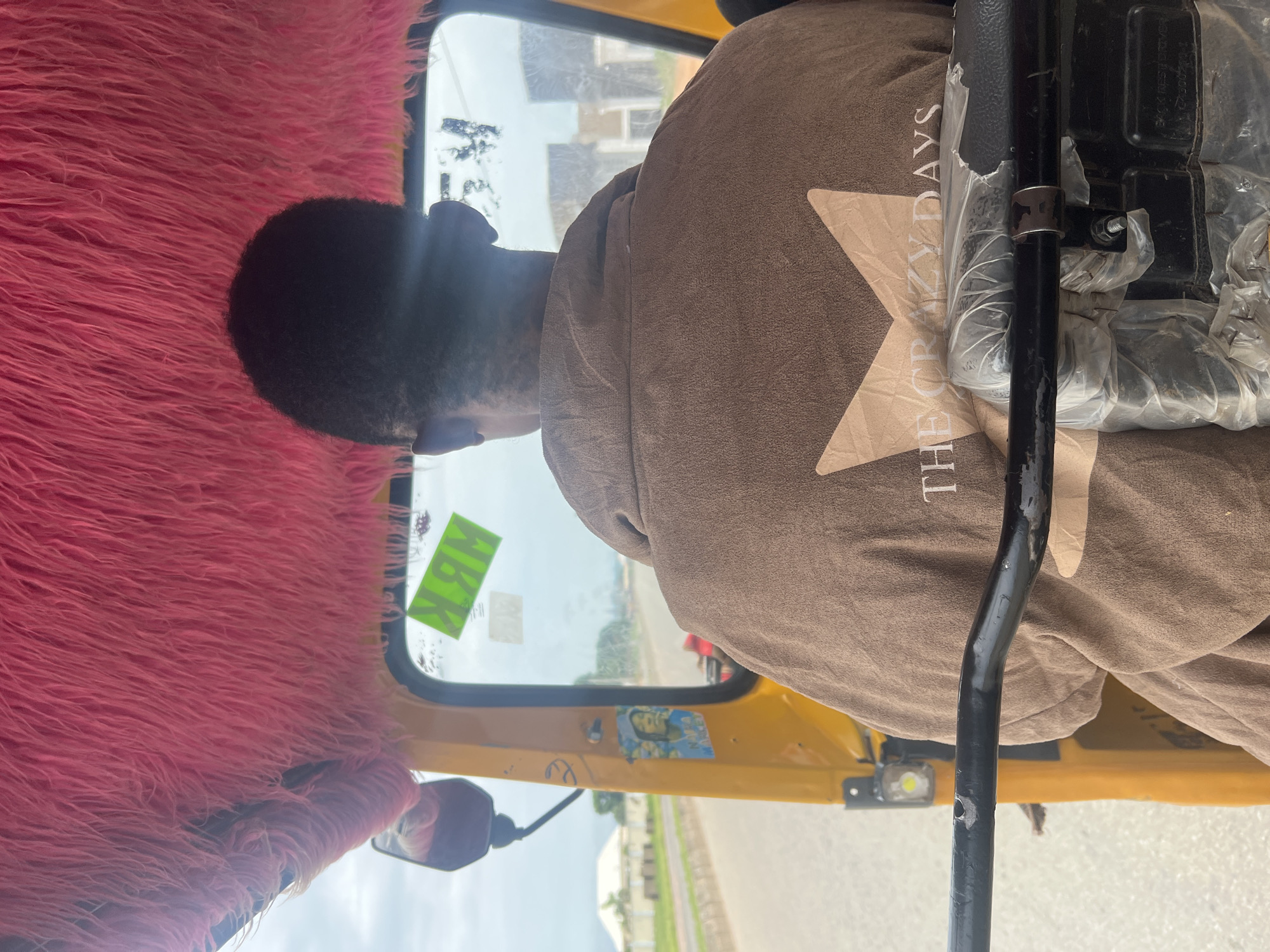 A person is shown from behind, driving a yellow auto-rickshaw with a fluffy pink interior, in Jalingo, Nigeria. The driver is wearing a brown jacket with a star design and the text "THE CRAZY DAYS" printed on the back. The auto-rickshaw is on a paved road, with buildings and houses visible in the background. The sky is overcast. There is a green sticker on the windshield that reads "NBM".