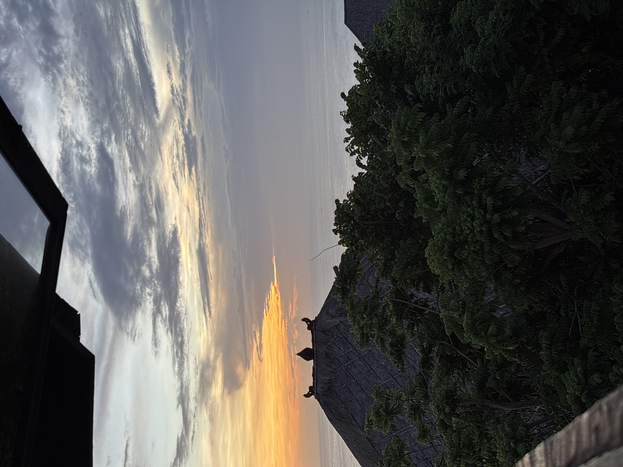 The image captures a sunset view, likely in Kuta Selatan, Indonesia, given the presence of traditional Balinese architectural elements. The sky is filled with clouds, transitioning from grey and white to orange and yellow near the horizon, indicating the sun's position. A traditional thatched roof structure with decorative statues is visible in the foreground, partially obscured by lush green foliage. The ocean can be seen in the background. A dark building corner is present on the left side of the image.