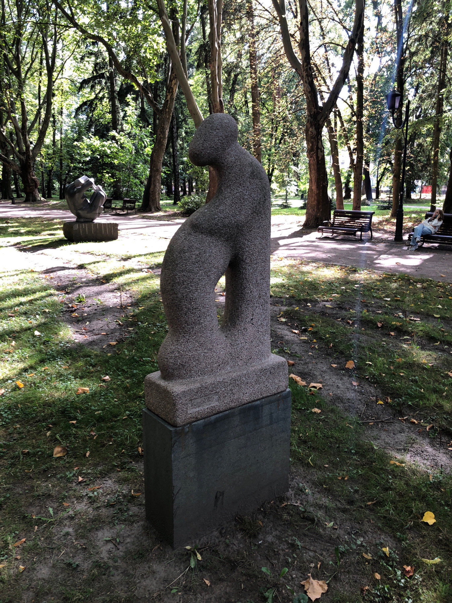 This is a medium shot of a stone sculpture in a park. The sculpture is abstract and appears to be made of granite. It is placed on a dark, rectangular pedestal. In the background, there are several trees with lush green foliage. Sunlight filters through the trees, creating dappled shadows on the ground. Further in the background, there are park benches with two individuals seated on one. The environment appears to be a public park in Lviv, Ukraine.