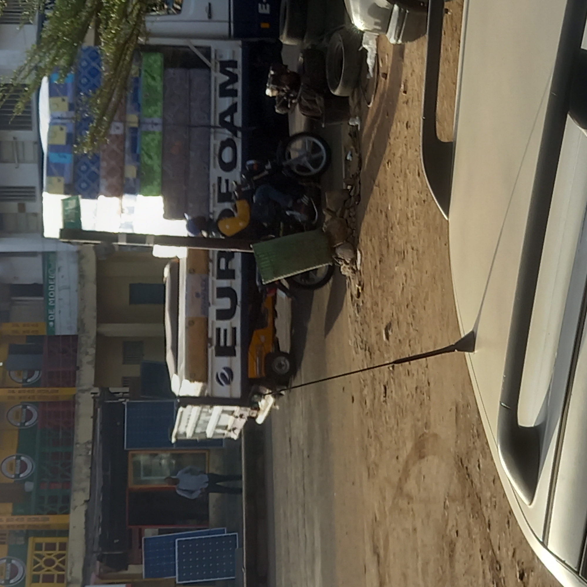 This image captures a vibrant street scene in Dadi Riba, Nigeria, under bright daylight. The main subject is a large white truck dominating the center-left, heavily loaded with a tall stack of multi-colored and patterned mattresses. The side of the truck clearly displays the word "EUROFOAM" in prominent dark letters, indicating its purpose as a mattress delivery vehicle.
To the right of the truck, a motorcycle is visible, with a person wearing blue jeans and a yellow top or helmet seated on it. Nearby, a smaller yellow utility vehicle, possibly a forklift, is partially seen. The ground is unpaved and dusty, characteristic of a local street, and there's some debris scattered around. The foreground reveals the side and antenna of a white car, suggesting the photograph was taken from within or beside it.
On the left side of the street, various commercial buildings or shopfronts are present. Stacks of colorful crates, possibly for beverages, are visible in front of one building. A sign above one of the shops reads "DE MODEL". Further down the street, another person in a light-colored shirt and dark trousers stands near a shop entrance. In the bottom left corner, two blue solar panels are visible, leaning against a surface. Lush green foliage from a tree is seen at the top, adding a natural element to the urban setting. The scene depicts a busy commercial thoroughfare, bustling with everyday activities like goods transportation and local commerce.