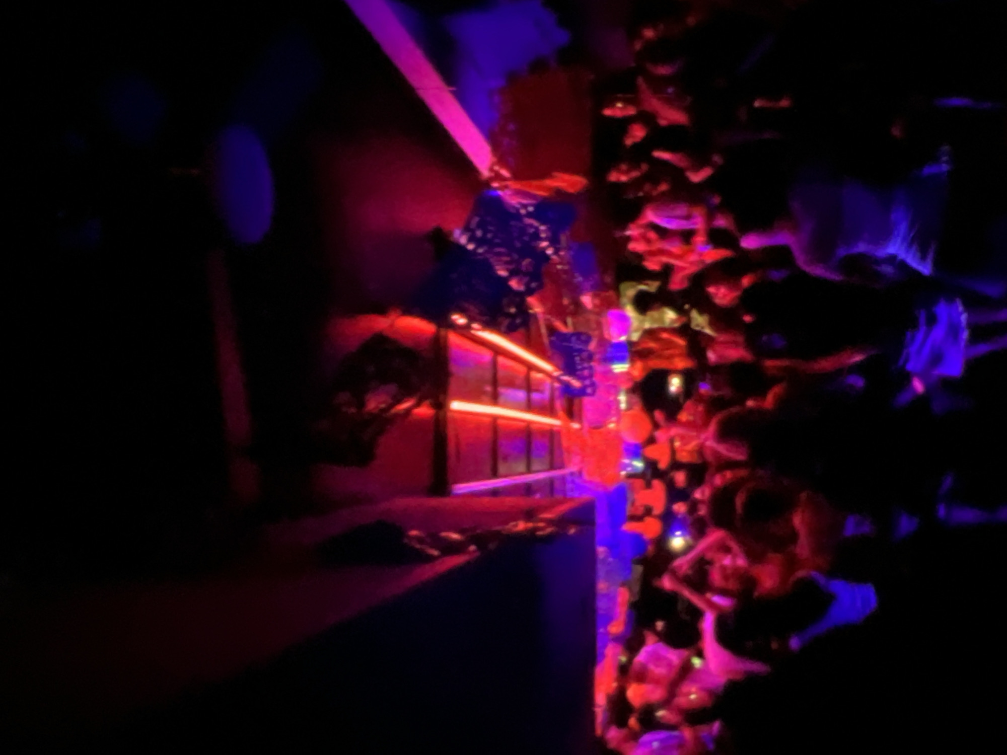 A group of people are dancing in a dark club with red and purple lights. The club appears to be in Lagos, Nigeria. There are several lights overhead, some of which are directed downwards to illuminate the dancers. The overall atmosphere is lively and energetic.