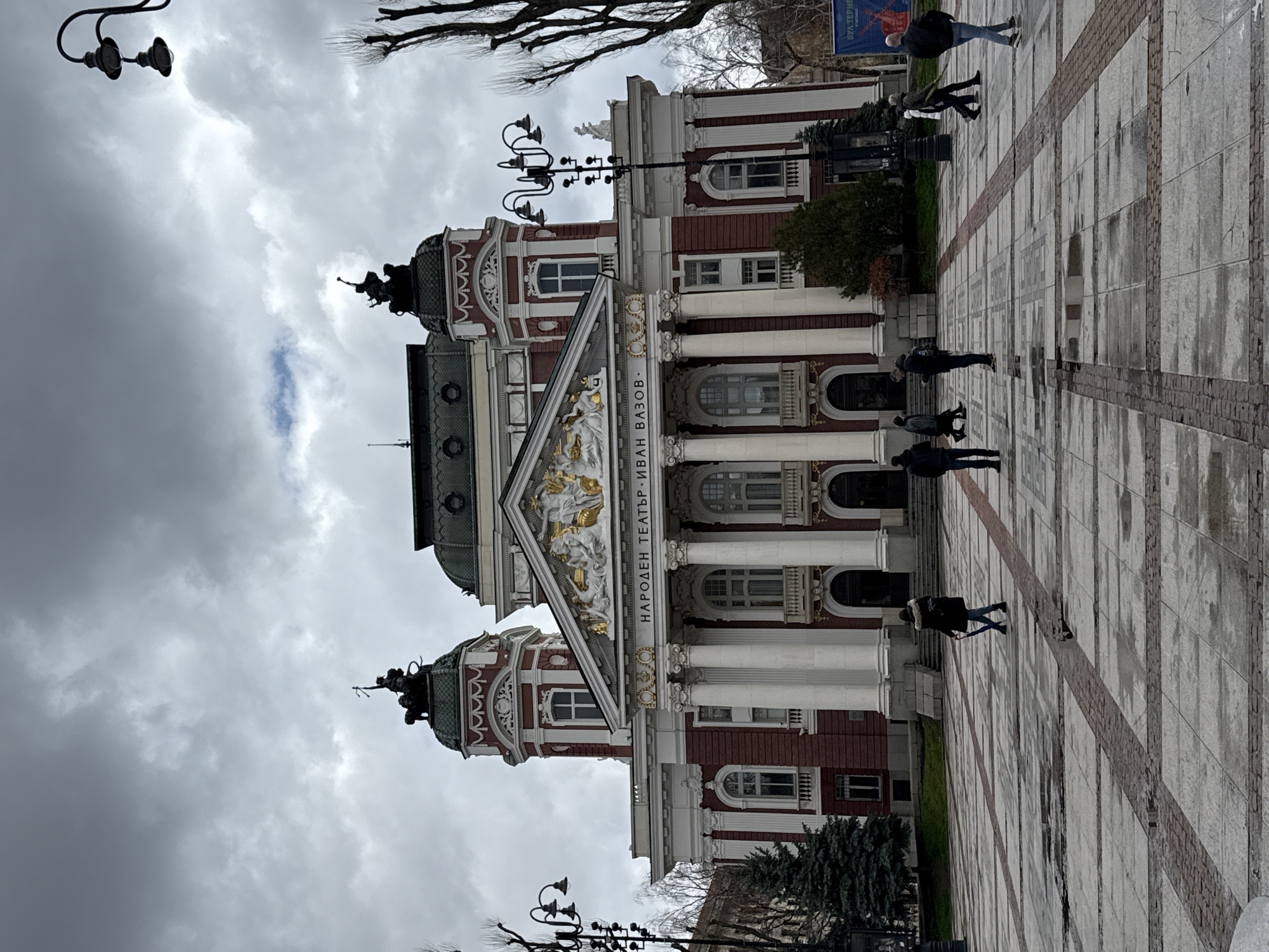 The image shows the Ivan Vazov National Theatre in Sofia, Bulgaria. The building is a grand structure with a red and white facade, columns, and sculptural details. The cloudy sky is visible in the background. In the foreground, people are walking across the paved square in front of the theatre. There are lamp posts and bare trees visible, along with a sign in the upper right corner.