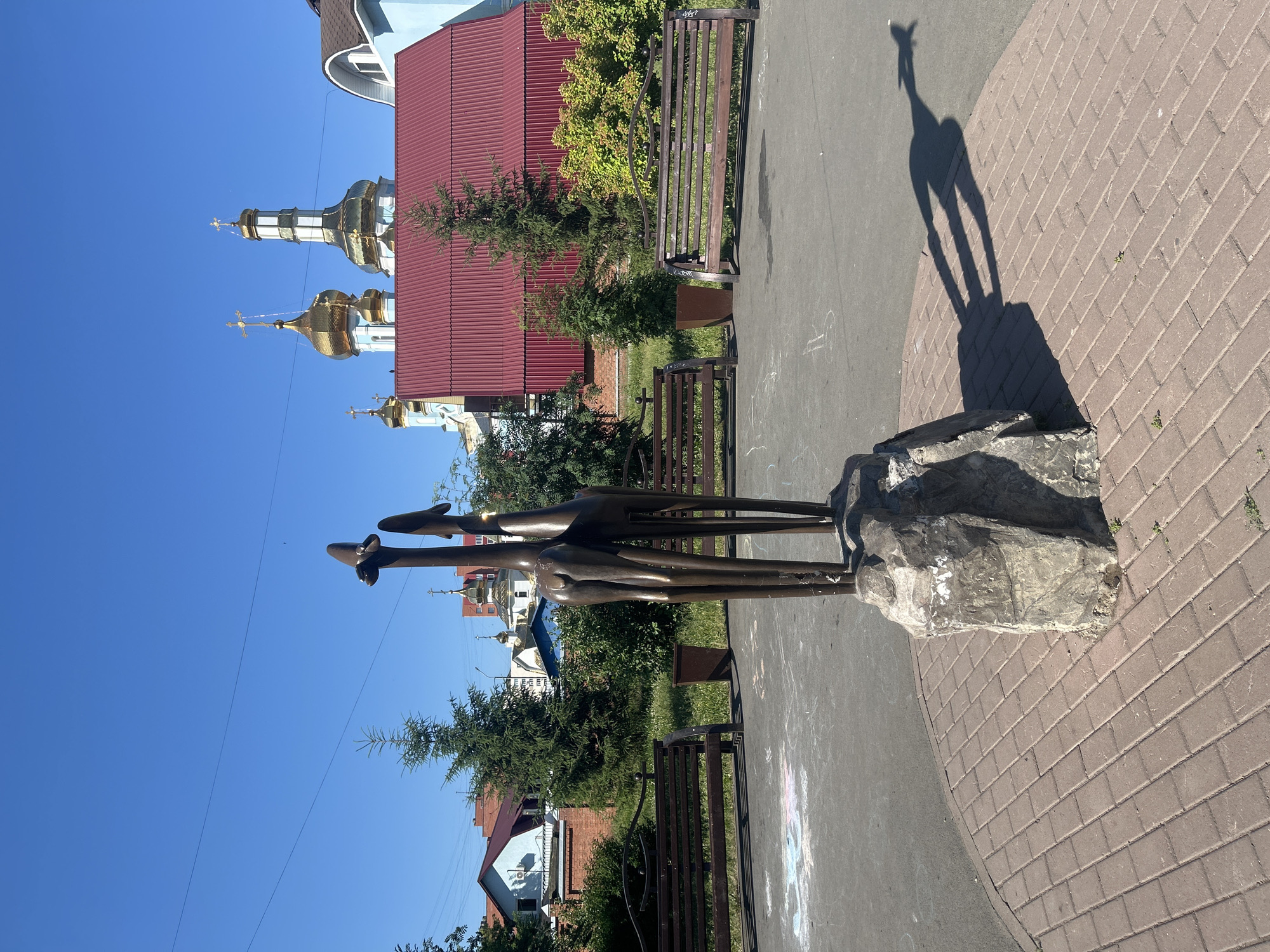 In Kemerovo, Russia, a public art installation featuring two abstract, metallic giraffe sculptures stands on a large rock base in a paved area. The sculptures are positioned in the foreground, casting long shadows onto the red brick paving. Behind the sculptures, a dark asphalt surface is visible, with traces of chalk drawings. Two dark wooden benches with metal frames are placed on either side of the paved area, and a dark trash receptacle is situated between them. Lush green bushes and trees provide a backdrop, with tall evergreen trees and smaller ornamental bushes. In the distance, a multi-domed church with golden cupolas and a red-roofed building are prominent against a clear blue sky. The overall setting appears to be a park or a public square.