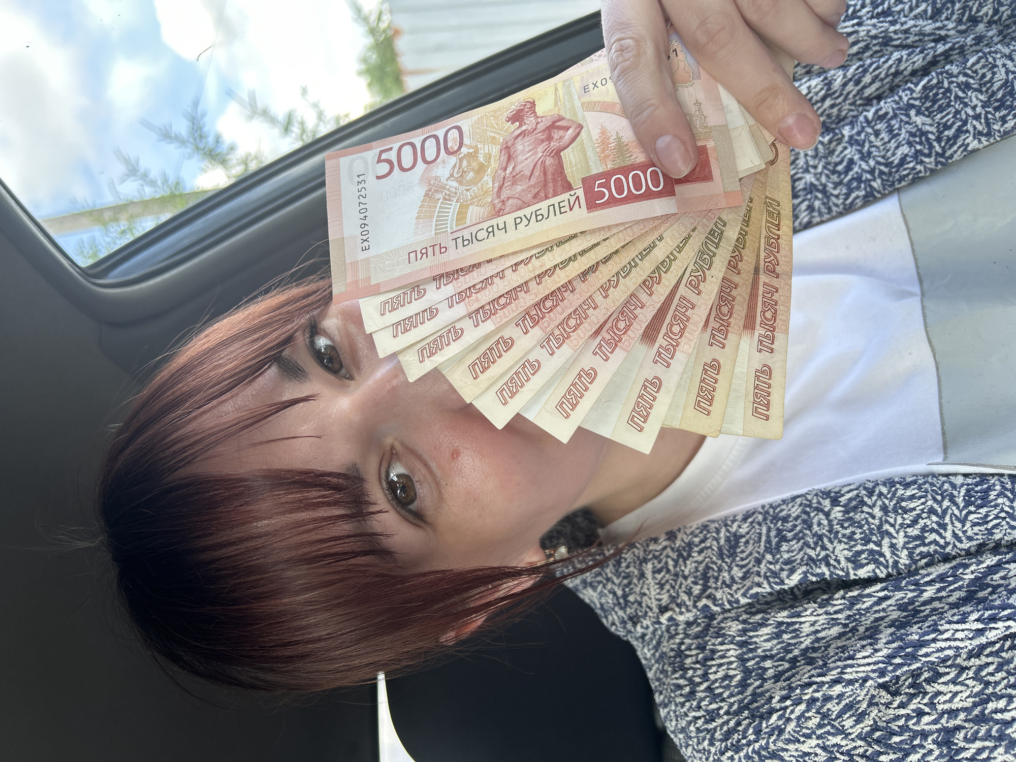 A close-up, slightly angled image captures a person, likely female, displaying a fan of Russian Ruble banknotes. Her reddish-brown hair with bangs partially covers her forehead, with one brown eye clearly visible looking towards the camera. She is wearing a white t-shirt layered under a blue and white patterned jacket or sweater. In her hand, she holds a fanned stack of at least seven 5000 Ruble banknotes. The top banknote clearly shows "5000" in large numerals, the phrase "ПЯТЬ ТЫСЯЧ РУБЛЕЙ" (FIVE THOUSAND RUBLES), and the serial number "EX094072531". A red-tinted depiction of a statue, likely the monument to Nikolay Muravyov-Amursky, is visible on the banknote. The background indicates an interior setting, possibly a car, with a window or sunroof revealing a cloudy sky and some tree branches, suggesting a daytime scene outdoors. The image conveys the act of holding or showcasing a significant amount of cash in Russia.