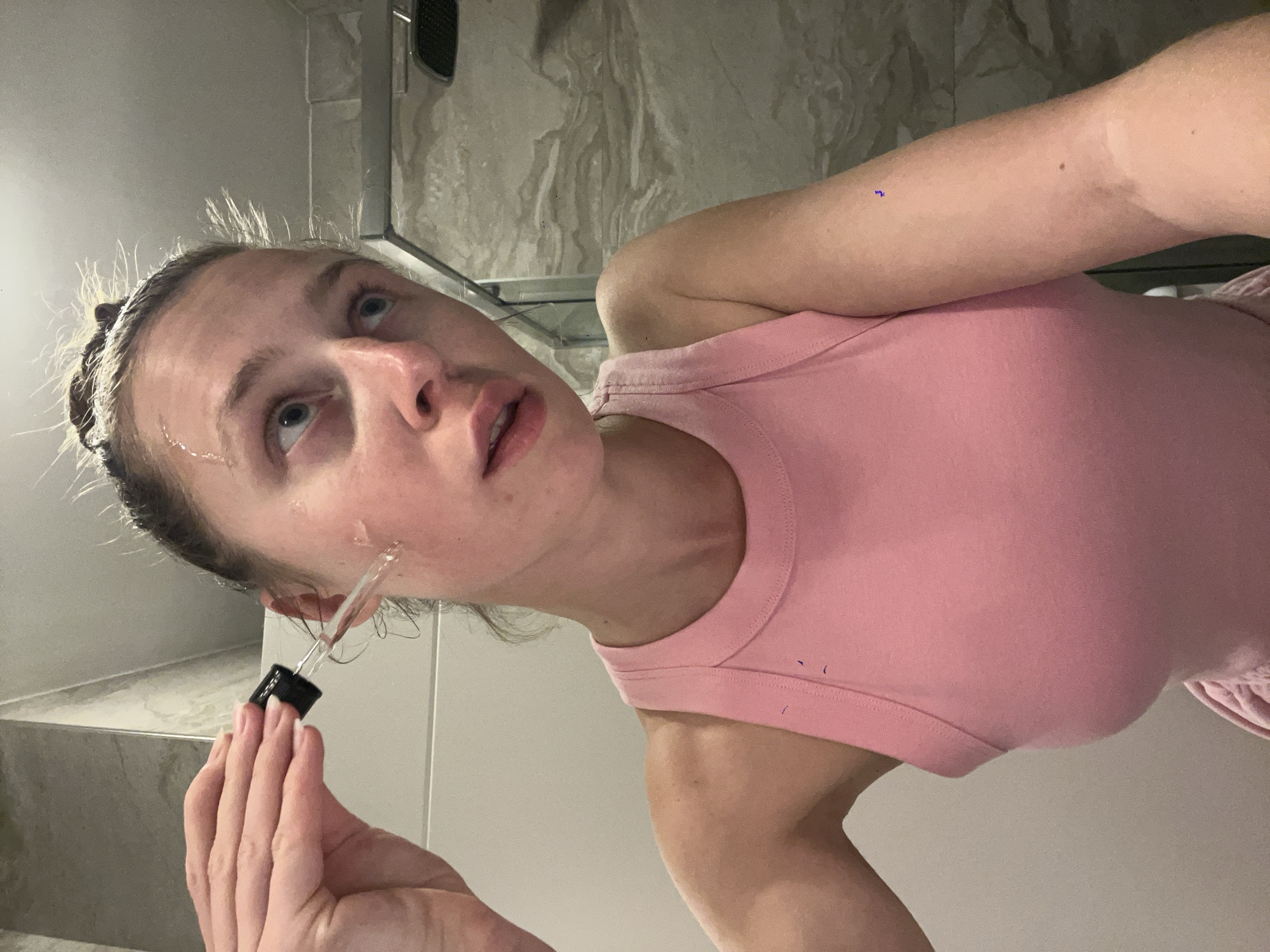 A woman is applying a liquid from a dropper to her face in a bathroom. She wears a pink tank top. The background includes marble and white tiles. The location is not discernible from the visual information provided.