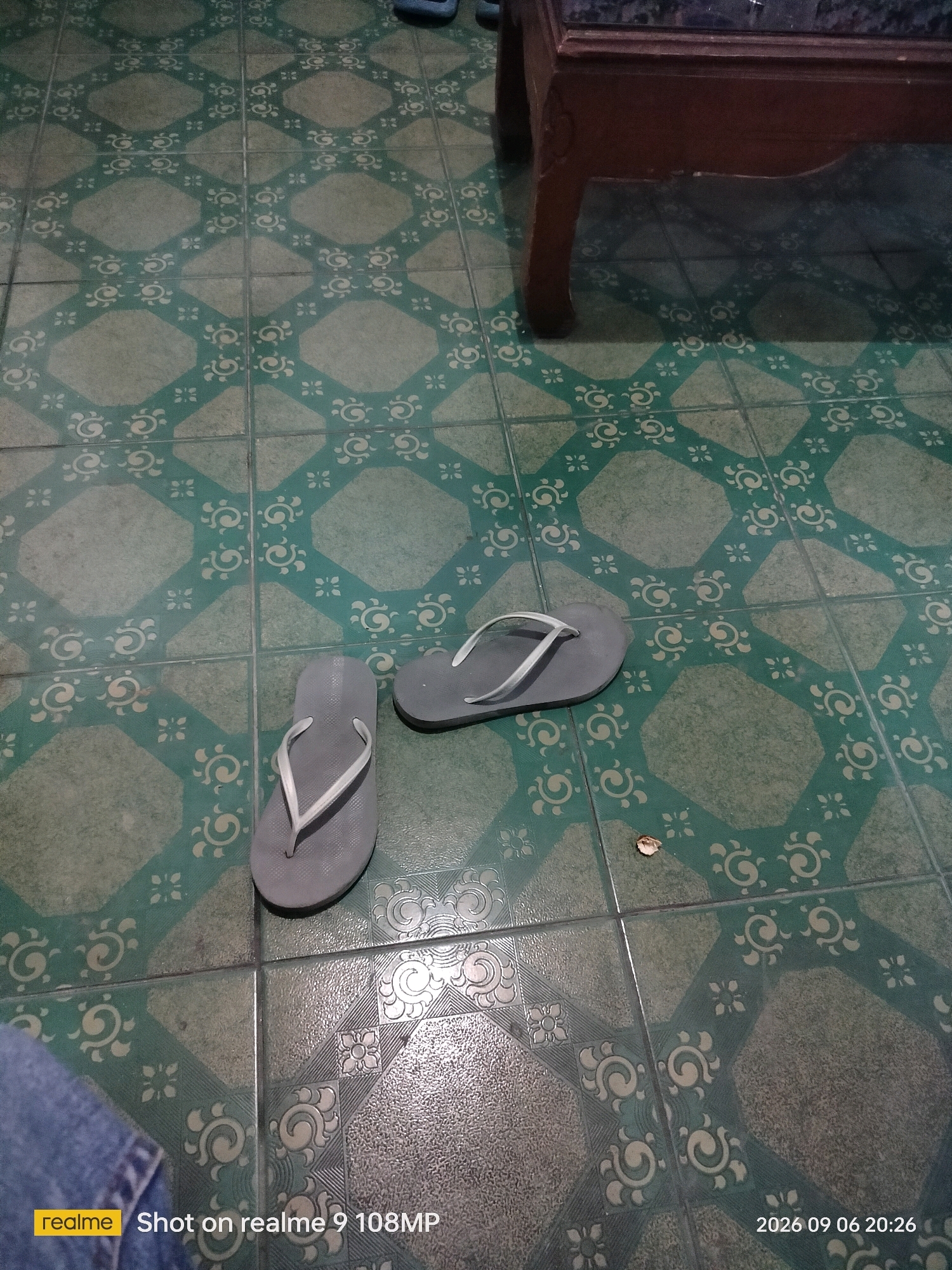 In an indoor setting, possibly a home in the City of Medan, Indonesia, a pair of gray flip-flops rests on a tiled floor. The floor is adorned with a repeating pattern of octagons and squares in green and white. The flip-flops are positioned near the center of the image, with one slightly in front of the other. To the right of the flip-flops, a small, dry leaf lies on the floor. In the upper right corner, a portion of a dark wooden table is visible. The lower left corner of the image shows a person's leg clad in blue denim jeans. A timestamp in the bottom right corner indicates the image was taken on September 6, 2026, at 8:26 PM.