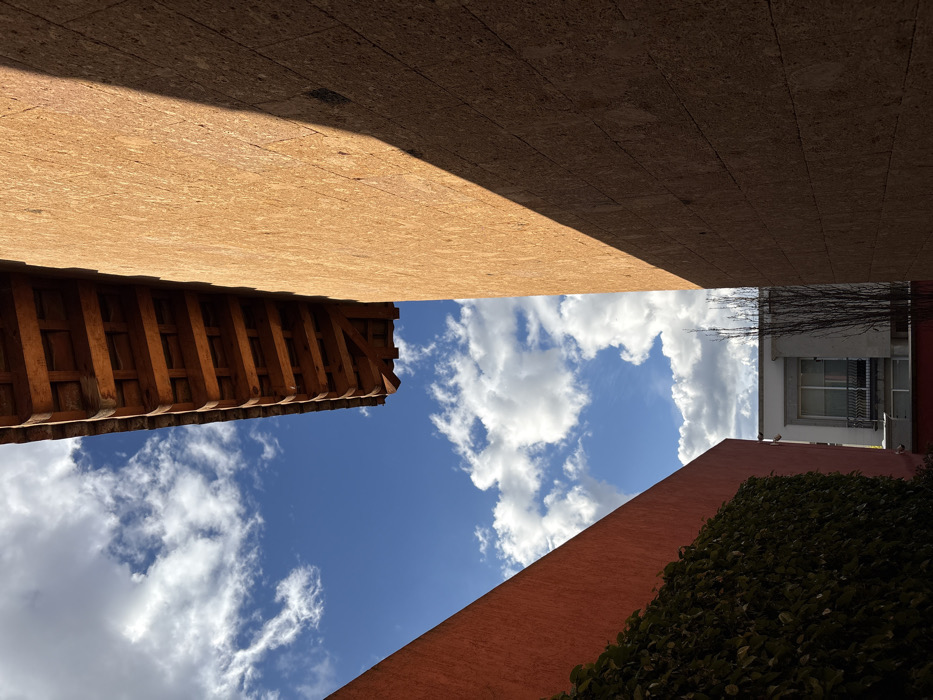 An upward-angled view showcases a vivid blue sky dotted with puffy white clouds, framed by building exteriors. A light brown, textured ceiling casts a shadow on one side. On the left, a wooden structure with regular beams projects into the frame. A reddish-orange wall and dark green foliage define the bottom corner, while a grey building facade with a window is visible on the right, completing the architectural enclosure.