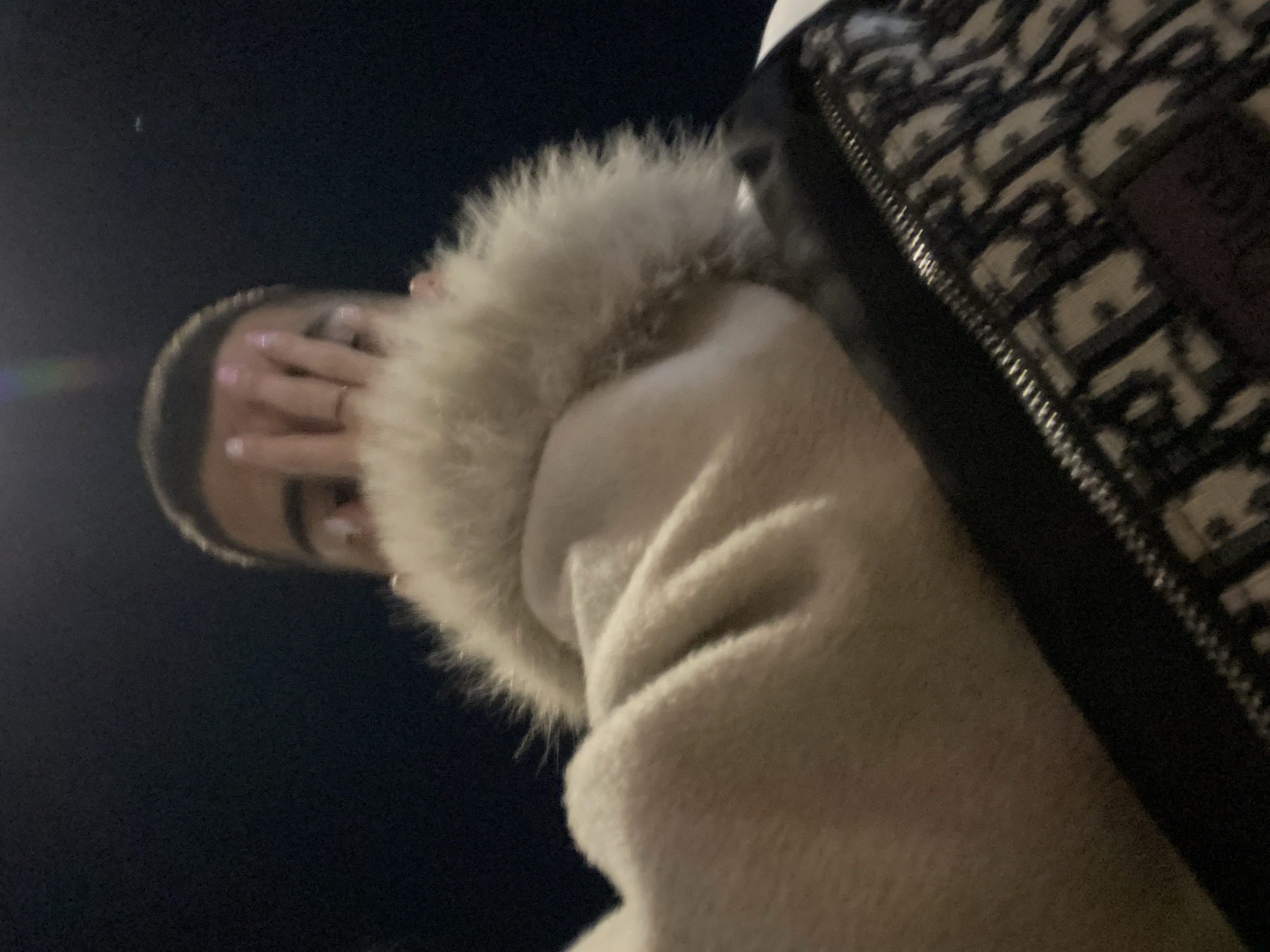 A low-angle shot depicts a person partly obscured by their coat and a patterned handbag. The person’s face is partially hidden behind their hand, revealing only a portion of their forehead and mouth. The background is dark, suggesting a night scene. The patterned handbag is situated to the right, with a visible zipper. The coat has a fur-lined hood, adding texture to the image. The exact location within Varna, Bulgaria, is not discernible from the visual information.