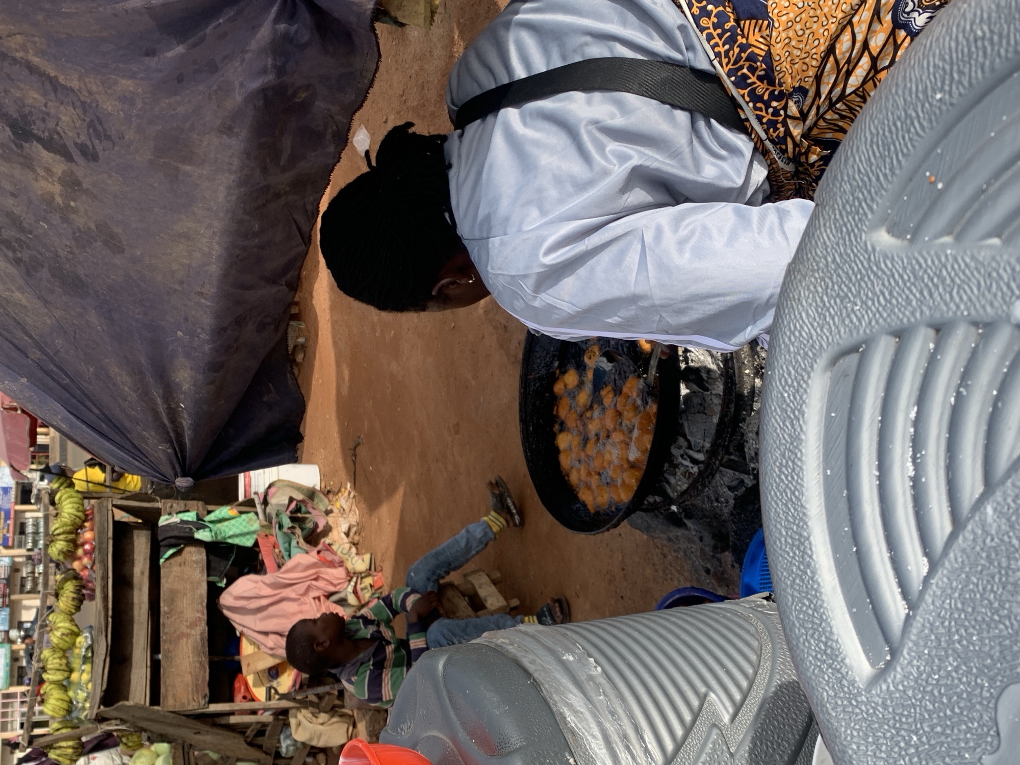 The image depicts an outdoor scene, likely a market, in the city of Gura Top, Nigeria. In the foreground, a large, ribbed grey plastic container is partially visible. Behind it, a woman with dark hair styled in a bun, wearing a light blue jacket and a vibrant, patterned skirt, is actively engaged in frying what appear to be fritters in a large black pan over an open fire. The pan sits on a makeshift stove made of charcoal. To the left of the frying woman, a young boy in a striped shirt and jeans sits on a small stool, observing the activity. Behind the boy, a wooden stall displays a variety of items, including what seem to be bunches of bananas and other produce, along with folded cloths and a red bucket. A large dark tarp is draped behind the stall and the frying station, providing some shade. The ground appears to be red earth, characteristic of the region.