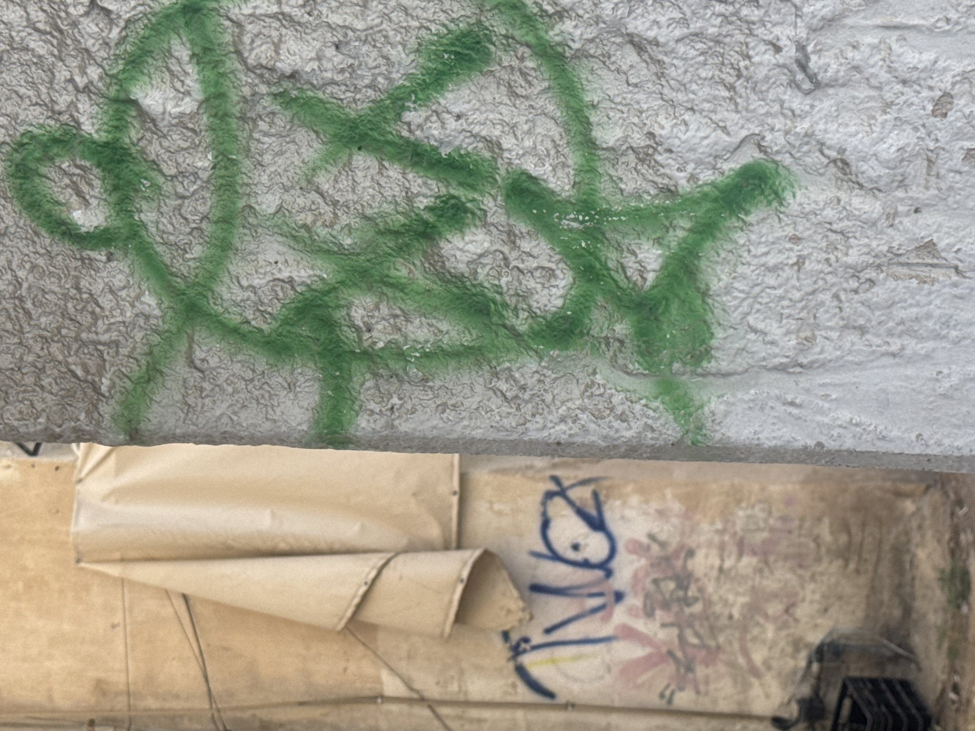 The image shows a section of an exterior wall in Givatayim, Israel. The upper part of the wall is rough and textured, with green graffiti covering much of the surface. Below the graffiti, there is a deteriorated and peeling section with remnants of an old sign. Blue graffiti markings are visible on the remaining section of the sign. The lower part of the image is out of focus, but hints at discarded objects and further deterioration of the area.