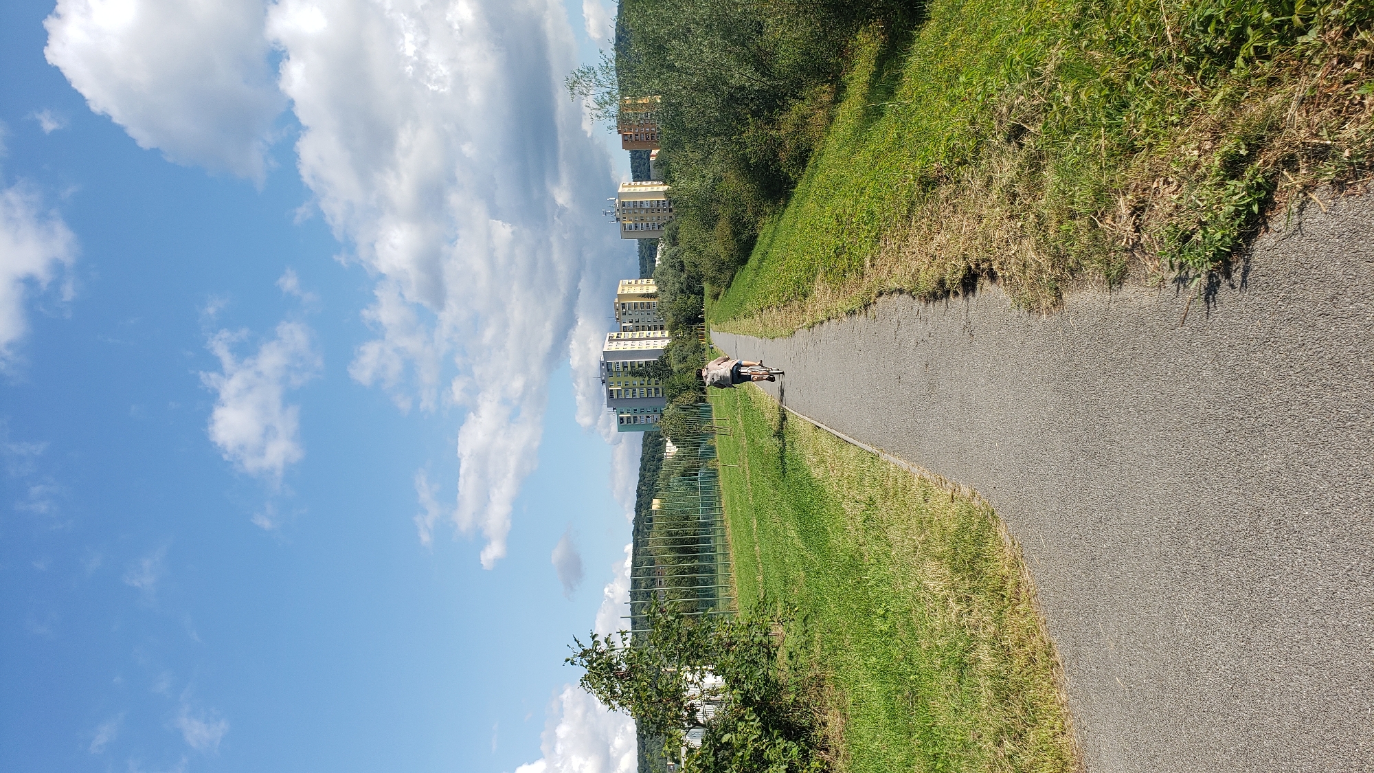 This is a photograph taken on a sunny day in the city of Prešov, Slovakia, looking along a paved pathway that winds through a grassy area. The pathway is bordered on the left by a mowed lawn and on the right by a more overgrown grassy embankment with some trees and bushes. In the middle ground, two people are cycling away from the camera on the pathway. Further in the distance, several multi-story apartment buildings are visible, set against a backdrop of rolling hills covered in trees. The sky above is a clear blue with scattered white cumulus clouds. The pathway appears to be part of a recreational area or a green belt within the city.