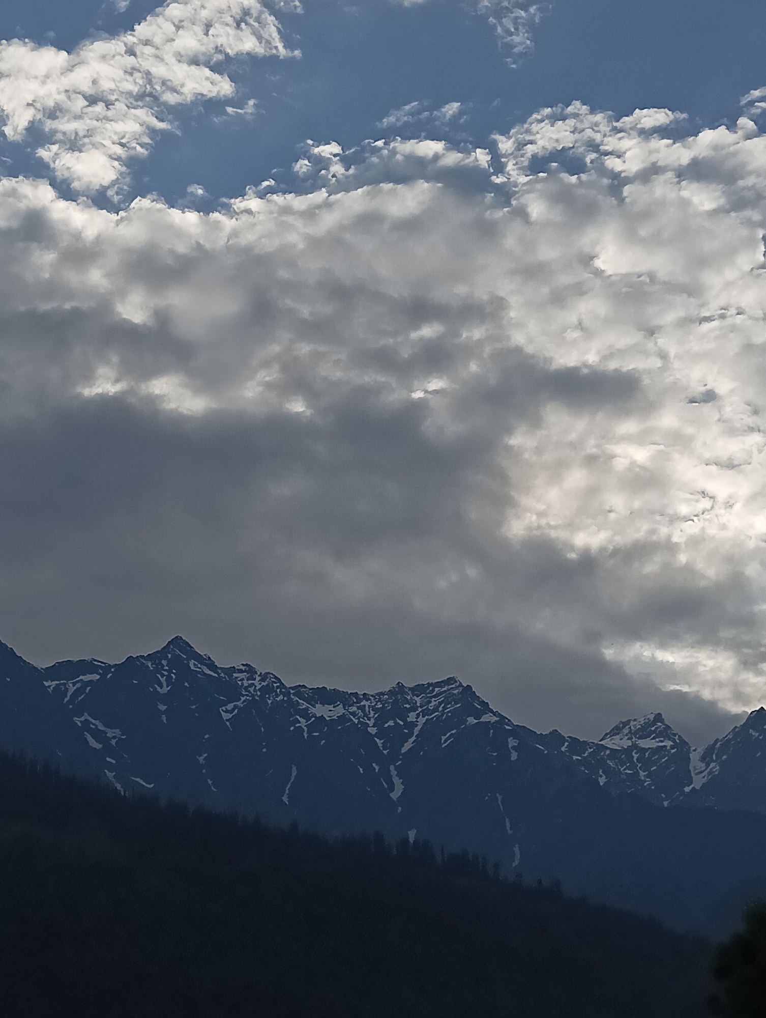 A dramatic sky filled with a mix of dark gray and white clouds looms over a jagged mountain range. The peaks of the mountains are dusted with snow, creating a stark contrast against the dark, rocky slopes. The foreground is a dark, shadowy expanse, possibly a forest or hillside, which transitions into the mountainous terrain. The lighting suggests either early morning or late evening, with the sun partially obscured by the clouds, casting an atmospheric glow. The image appears to be a landscape photograph taken from a distance, capturing the grandeur of the natural environment.